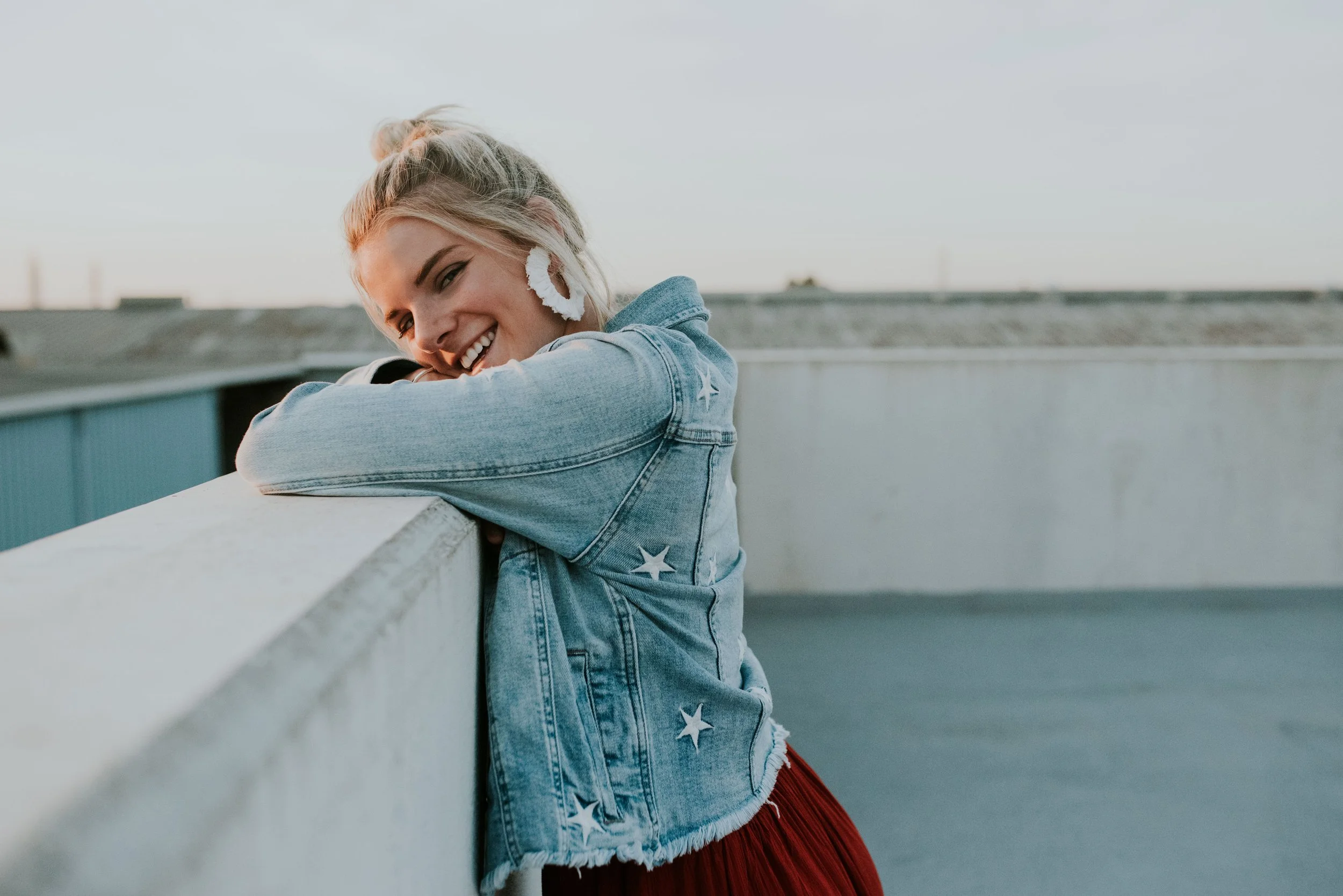 A young woman with blonde hair styled in a messy bun, wearing large white earrings and a denim jacket with star patches, smiling and leaning on a white concrete barrier on a rooftop with a cloudy sky.