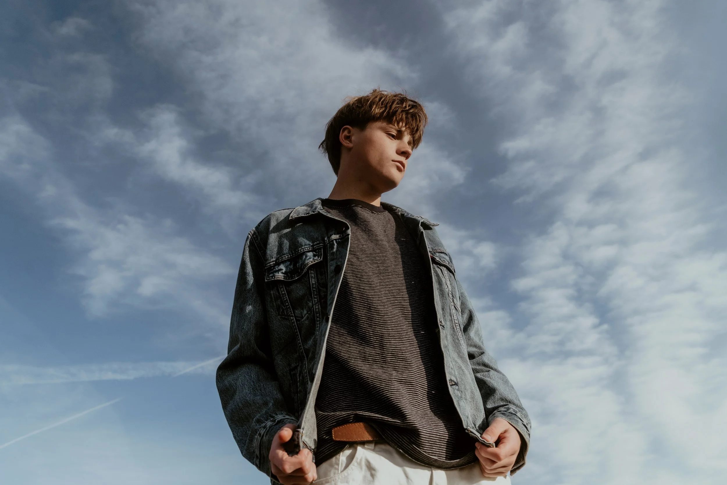 Young man with brown hair wearing a denim jacket and striped shirt, standing outdoors against a cloudy sky.