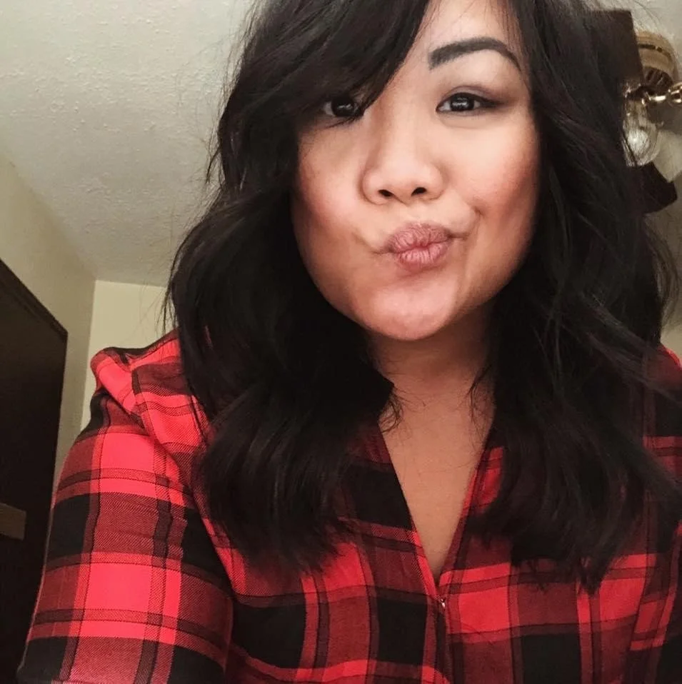 A woman with dark, wavy hair taking a selfie, making a puckered face, wearing a red and black plaid shirt, in a room with a beige wall and a ceiling light fixture.