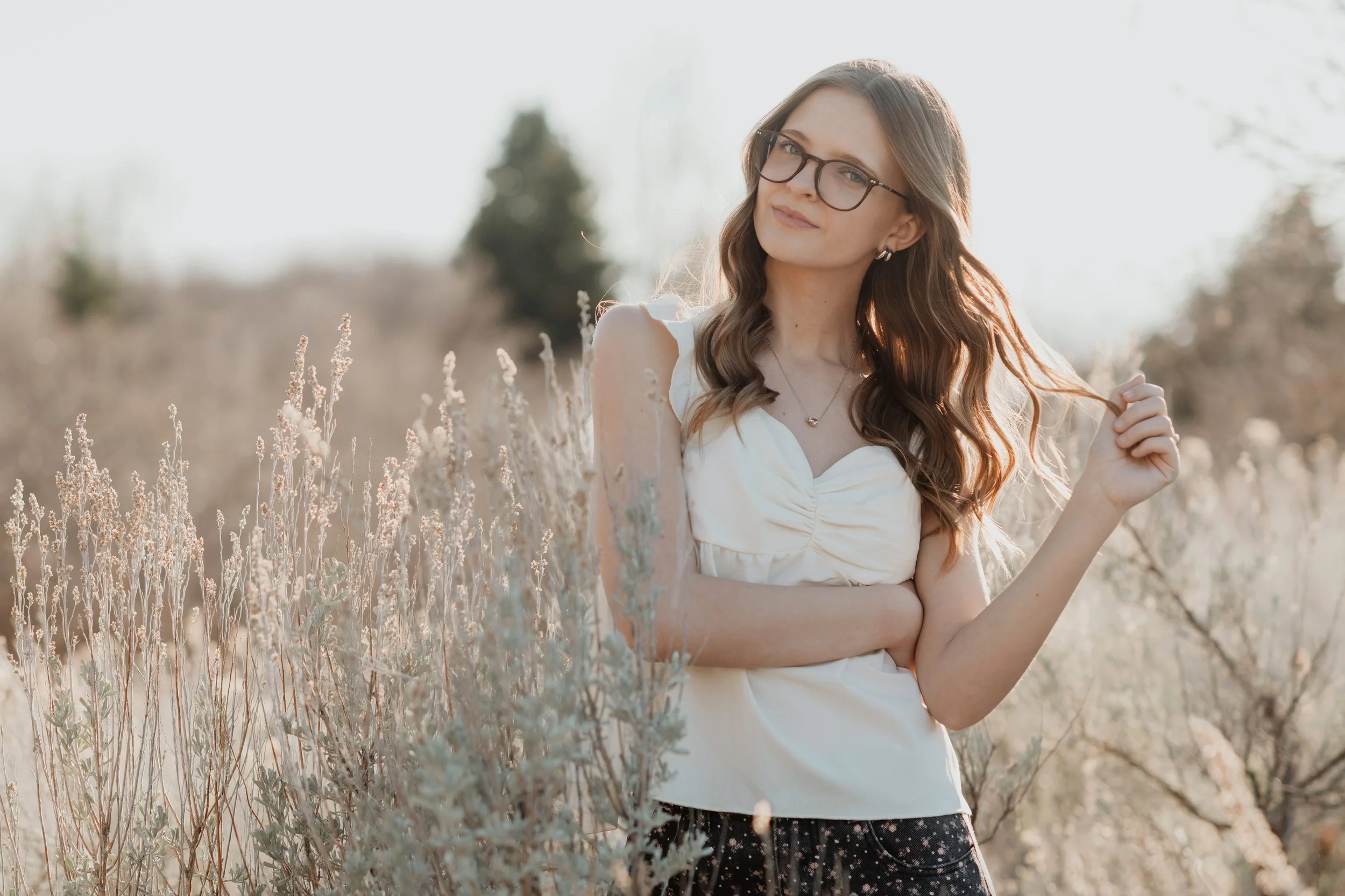 A young woman with long wavy brown hair wearing glasses, a white sleeveless top, and a necklace, standing in a field of tall, dry plants during daylight, with trees and a cloudy sky in the background.