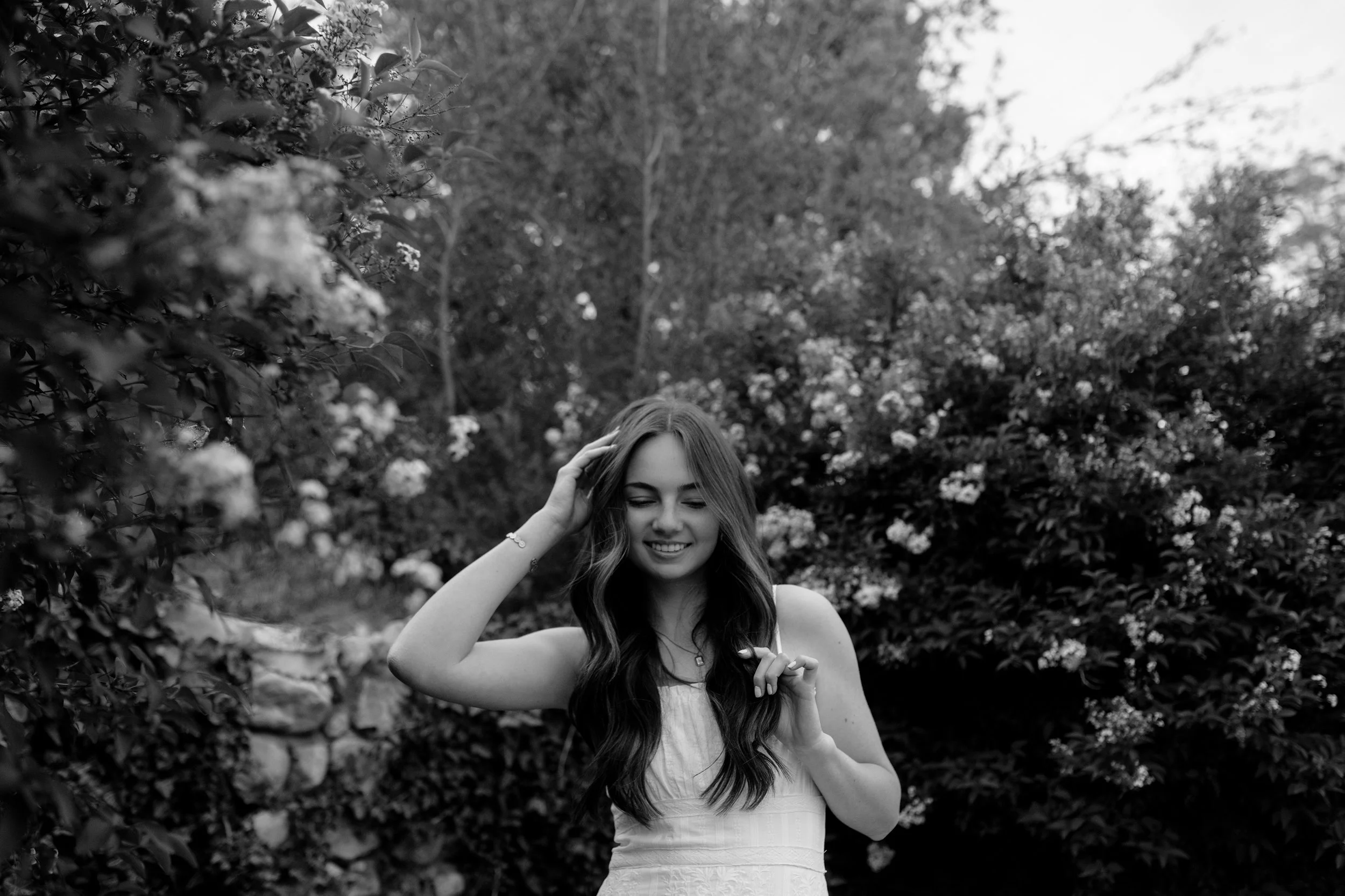 A smiling young woman with long hair standing outdoors among bushes and trees, touching her hair with one hand.