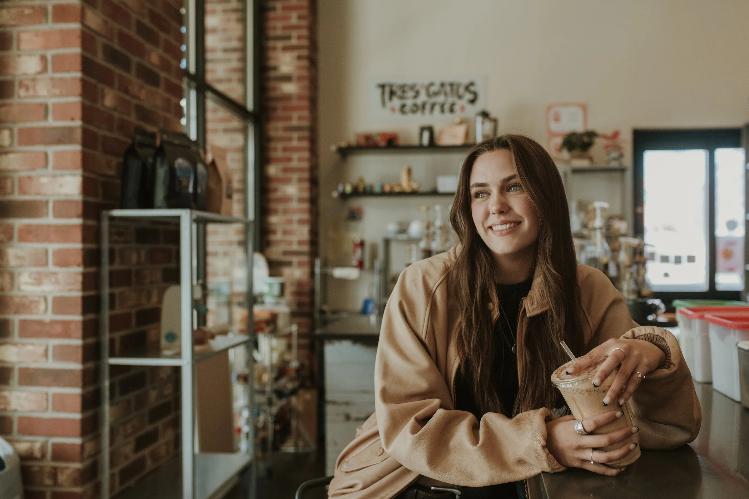 A young woman with long brown hair and a tan jacket sitting inside a coffee shop holding a plastic cup with a drink and smiling.