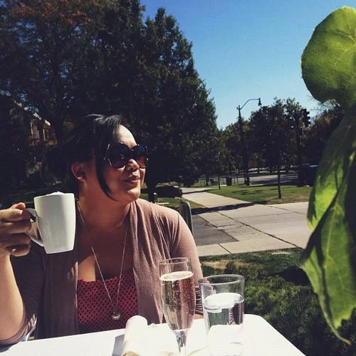 Woman sitting outdoors holding a coffee mug, wearing sunglasses, with a glass of water and a glass of champagne on the table in front of her; sunny day with trees and a sidewalk in the background.