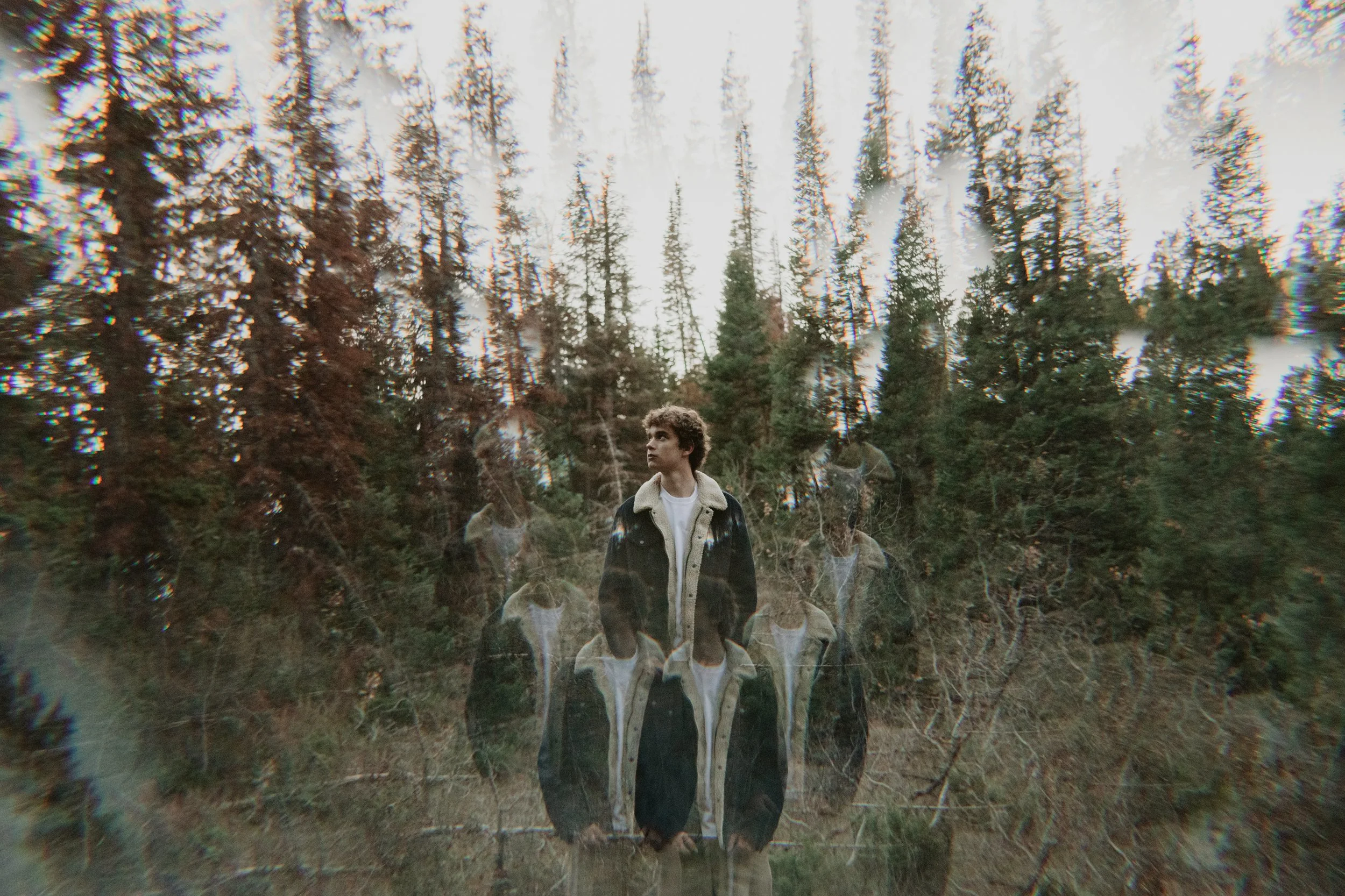 A young man with curly hair wearing a jacket stands among trees in a wooded area with many distorted and overlapping images, creating a surreal, ghost-like effect.