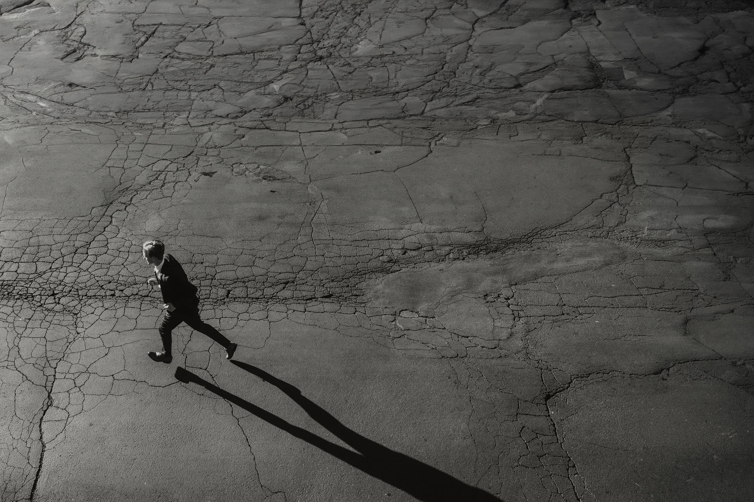 A man walking on a cracked asphalt road casting a long shadow, with the image taken from above in black and white.