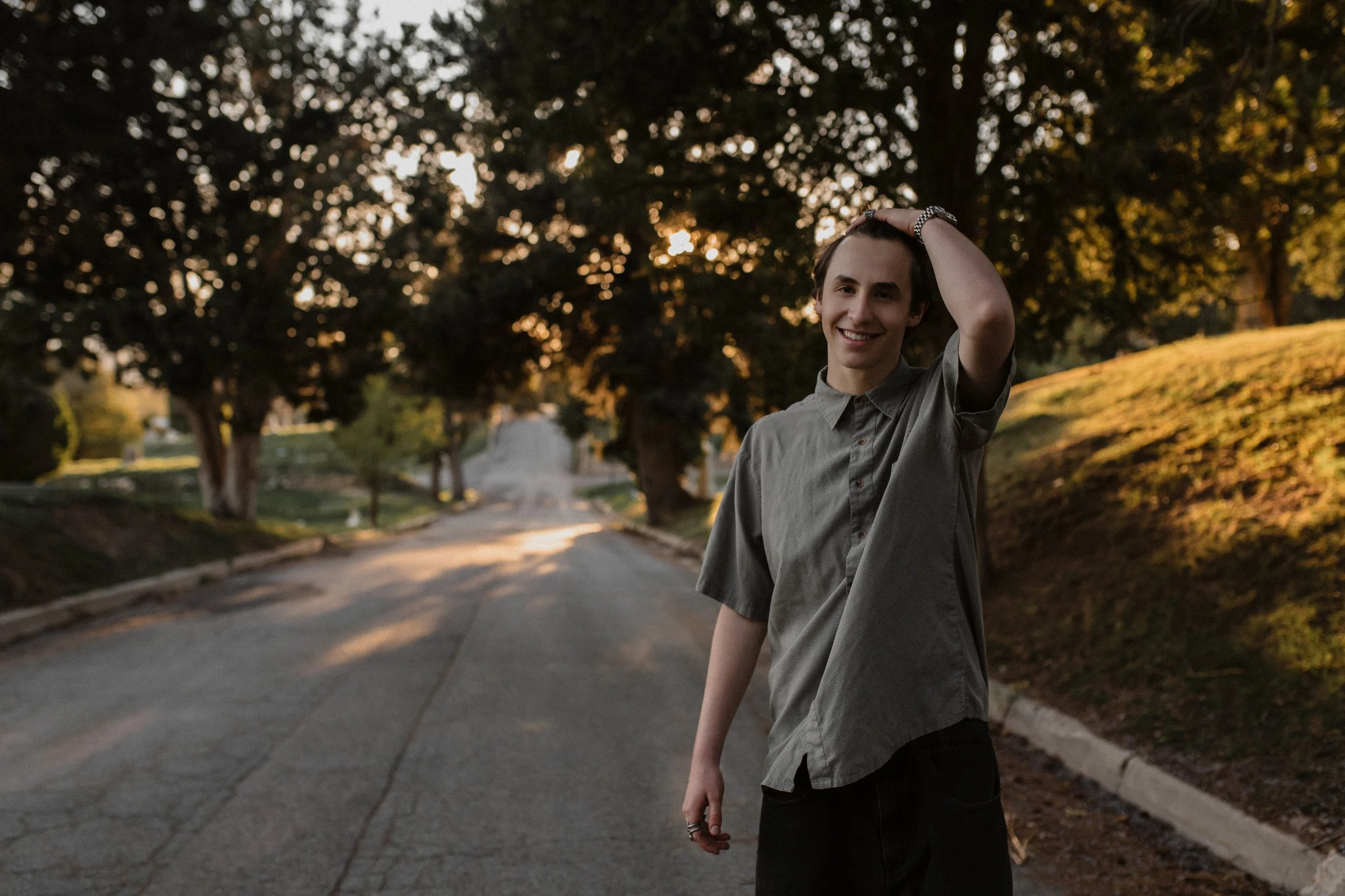 A young man with dark hair smiling and touching his hair, standing outdoors on a street lined with trees during sunset.