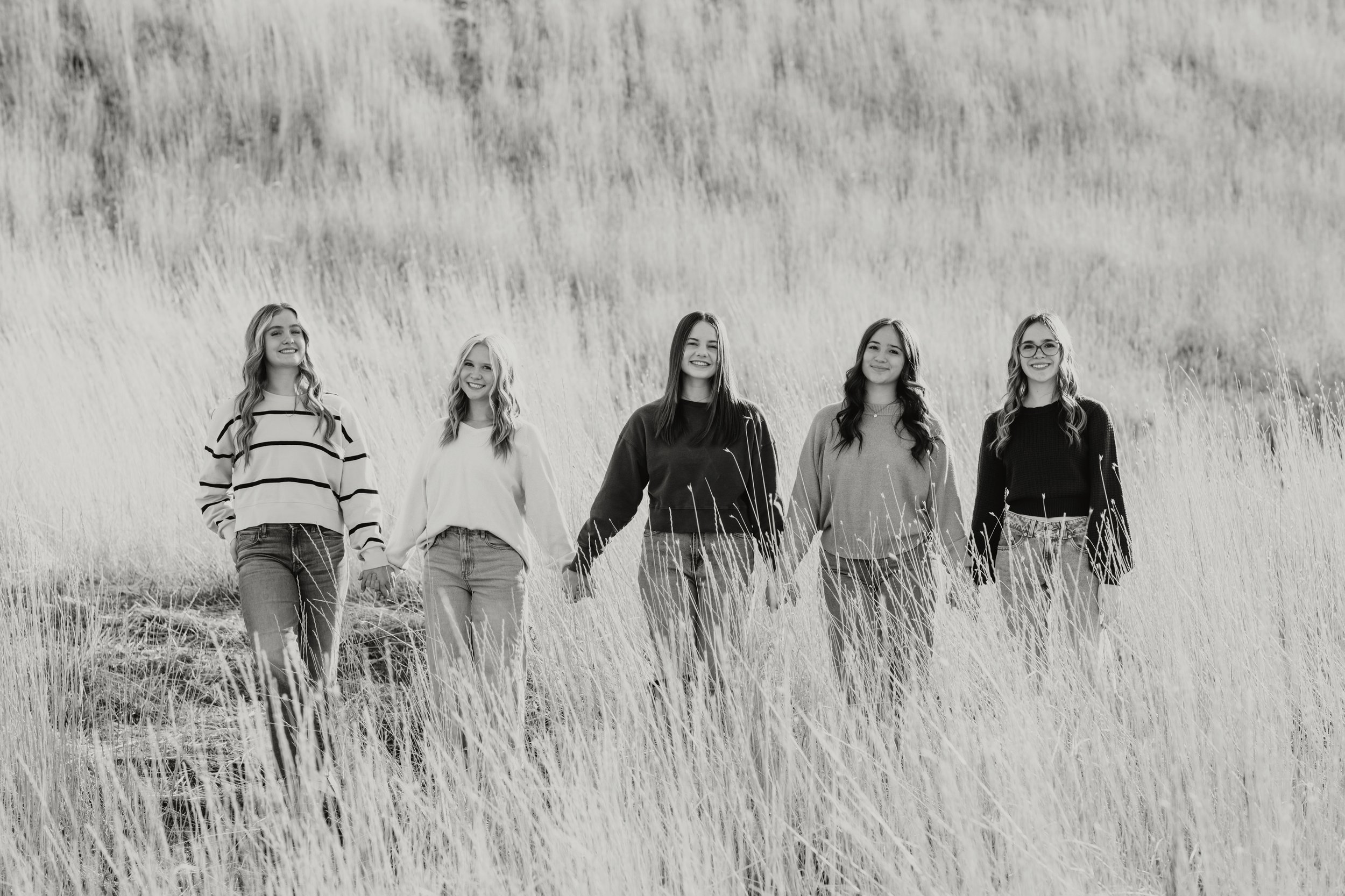 Five young women walking together in a grassy field, smiling, with a hillside in the background, in black and white.