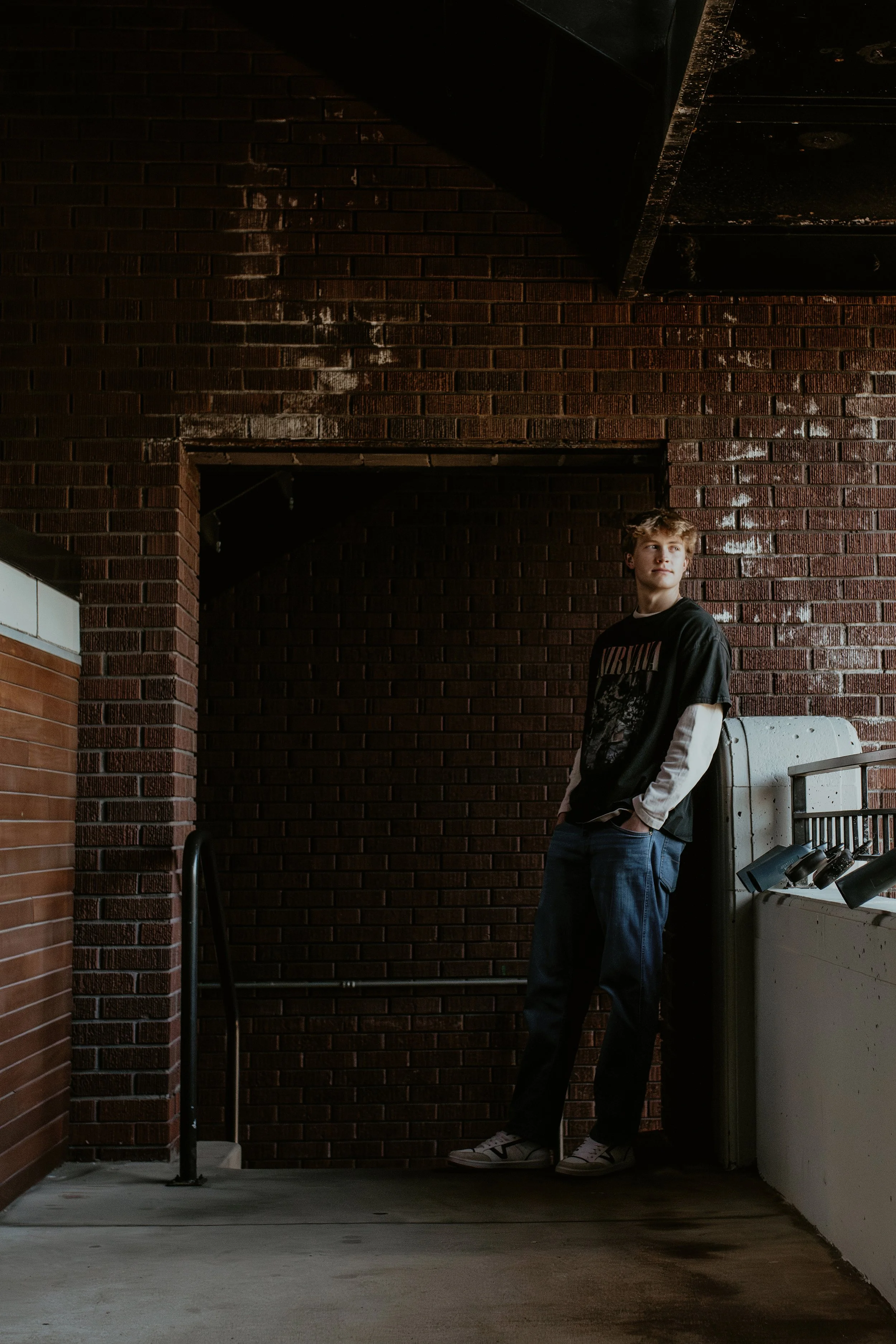 A young man with curly hair is standing against a red brick wall near a white washing machine. He is wearing a black Nirvana T-shirt over a long sleeve white shirt, blue jeans, and white sneakers. The background shows a dark overhead structure and a 