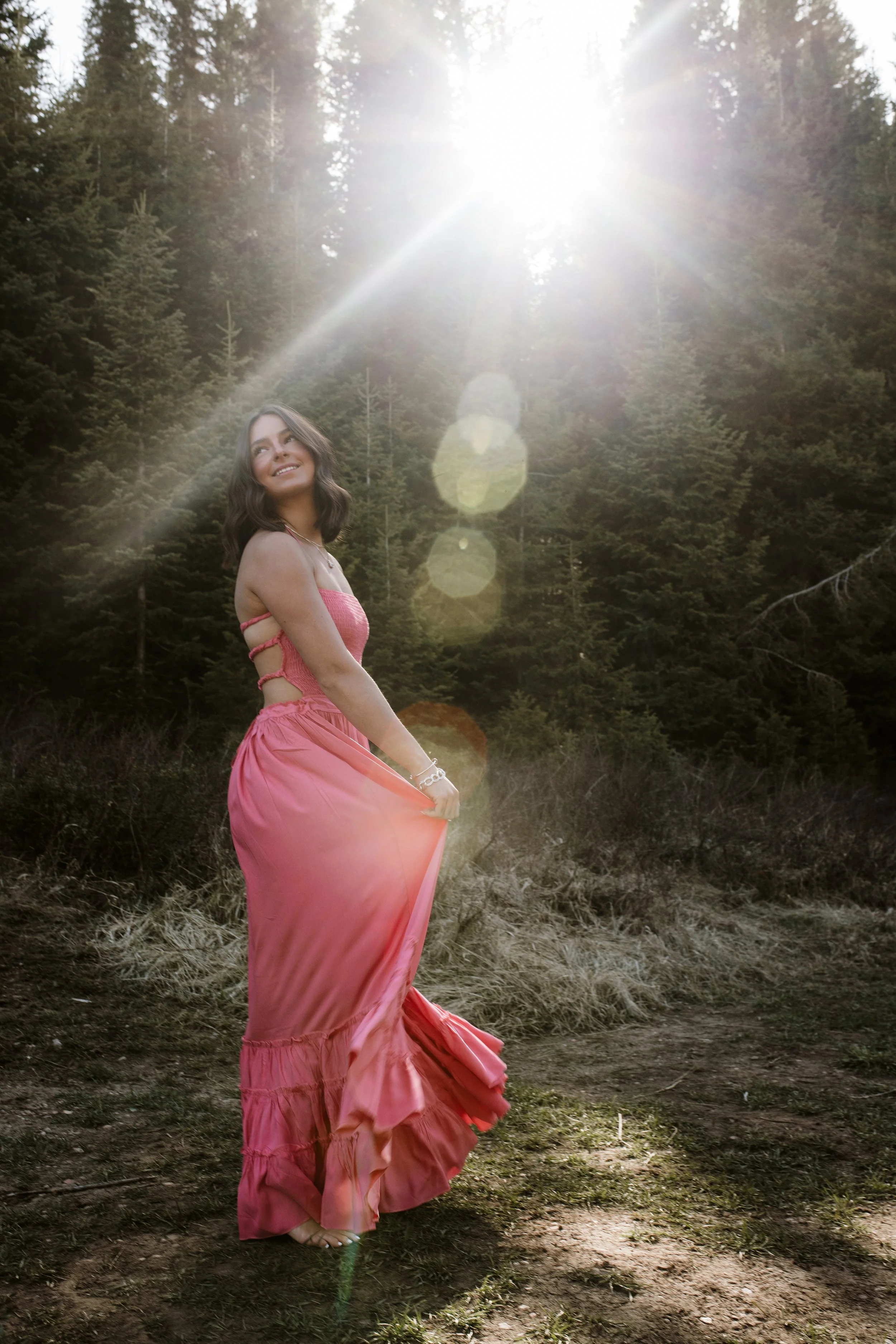 A woman in a flowing pink dress standing outdoors in a forest, with sunlight streaming behind her creating a lens flare.