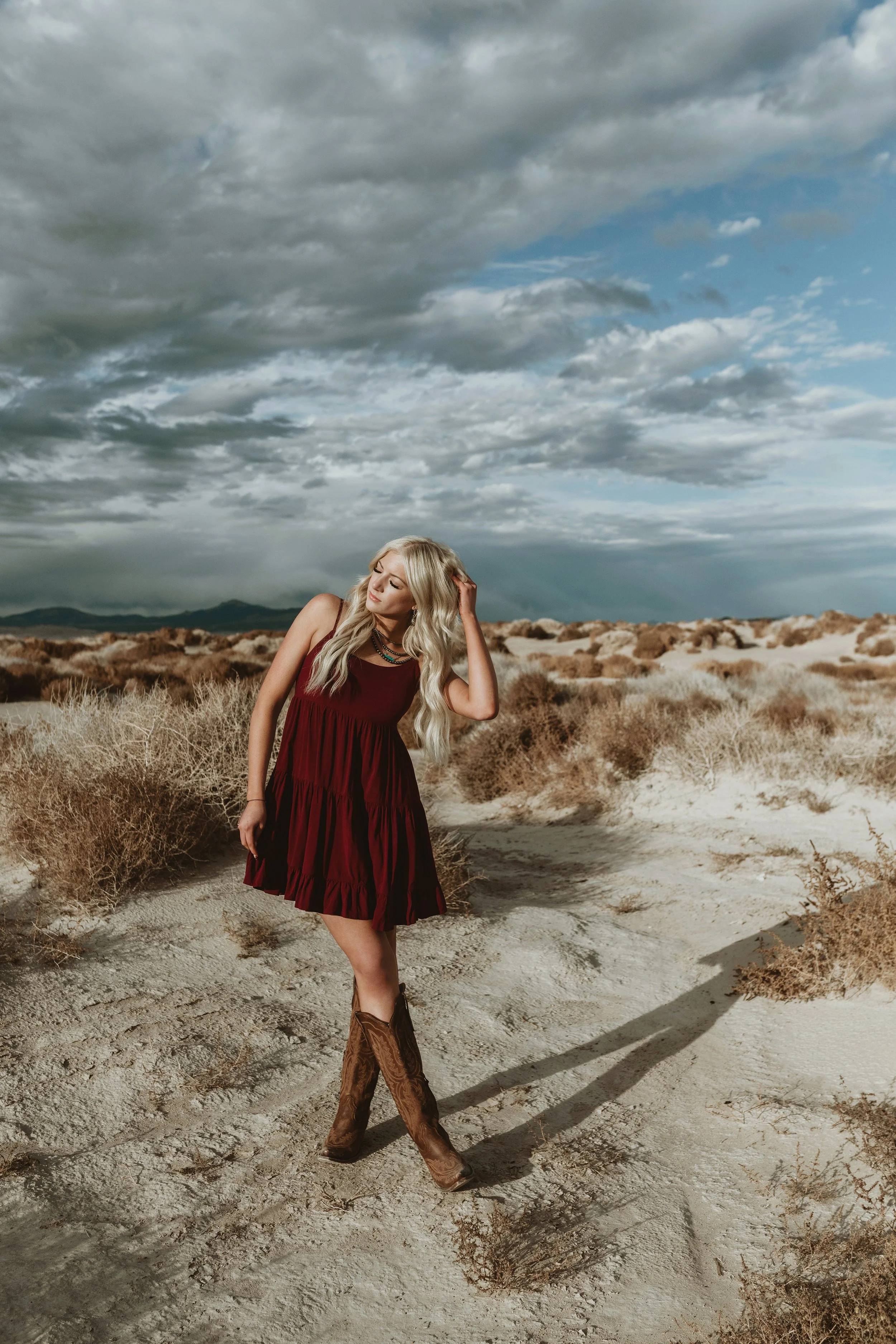 Blonde woman in a burgundy dress and cowboy boots standing on a sandy, desert landscape with sparse bushes, under a cloudy sky.