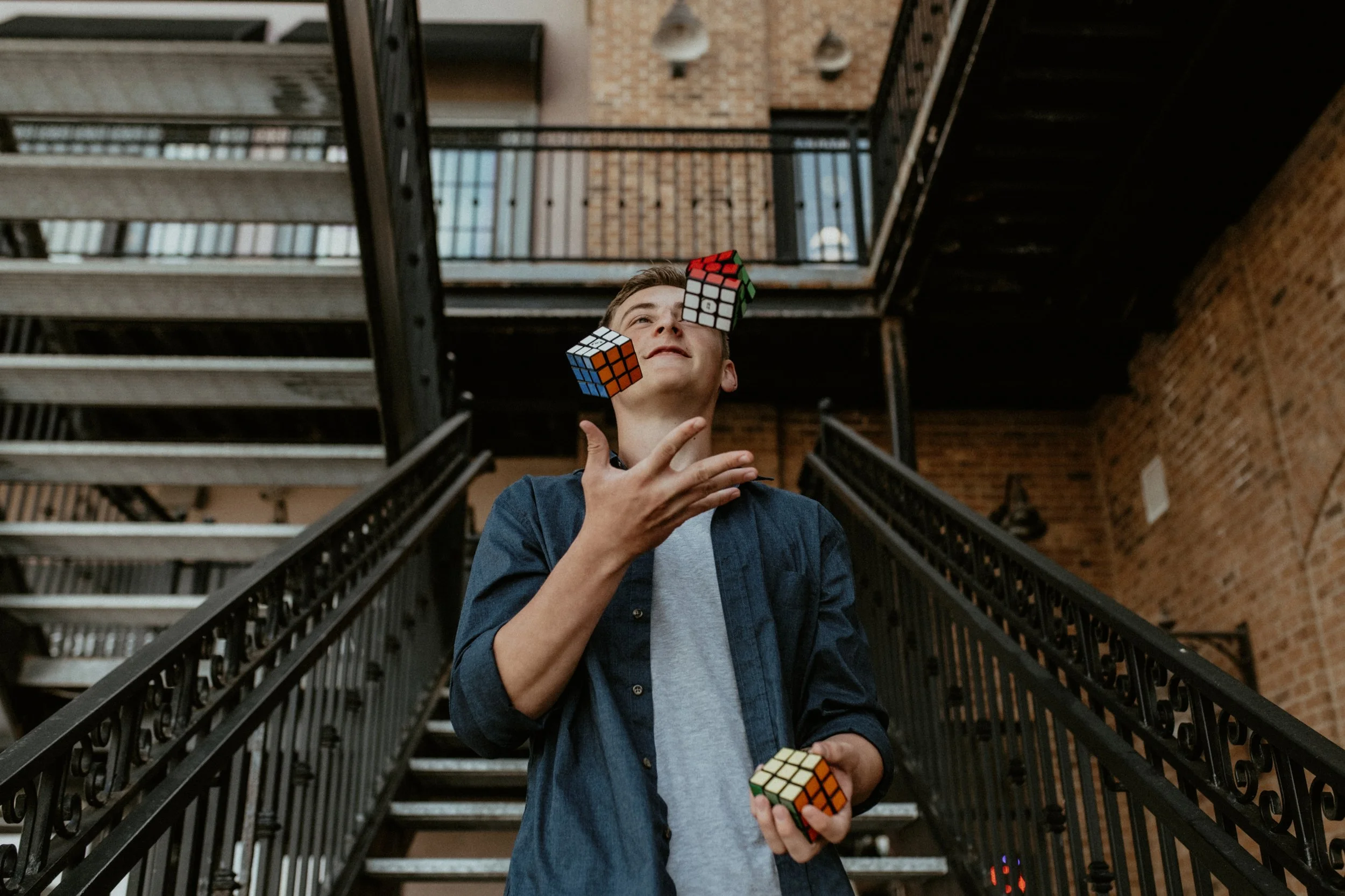 A young man holding multiple Rubik's cubes on a staircase in an industrial-style building with brick walls and black metal railings.