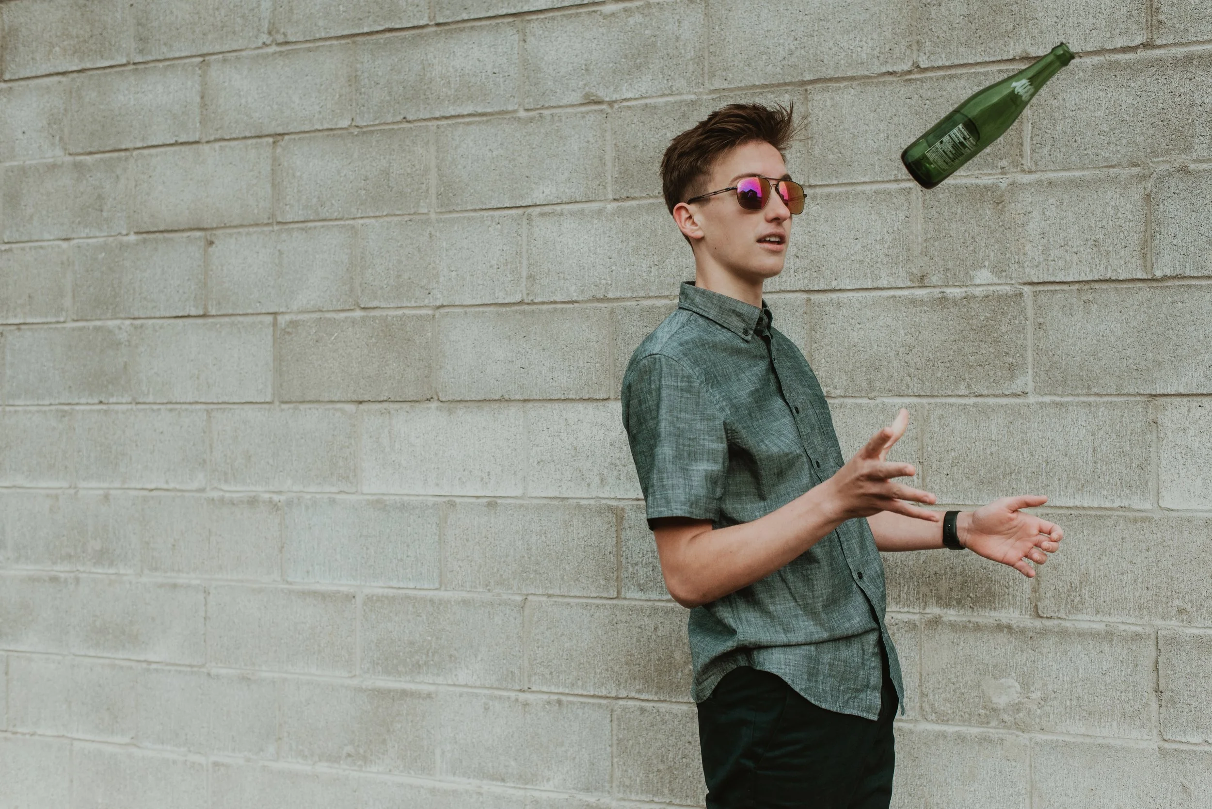 A young man wearing sunglasses and a short-sleeved button-up shirt stands against a beige brick wall, looking at a green glass bottle that appears to be floating or falling.
