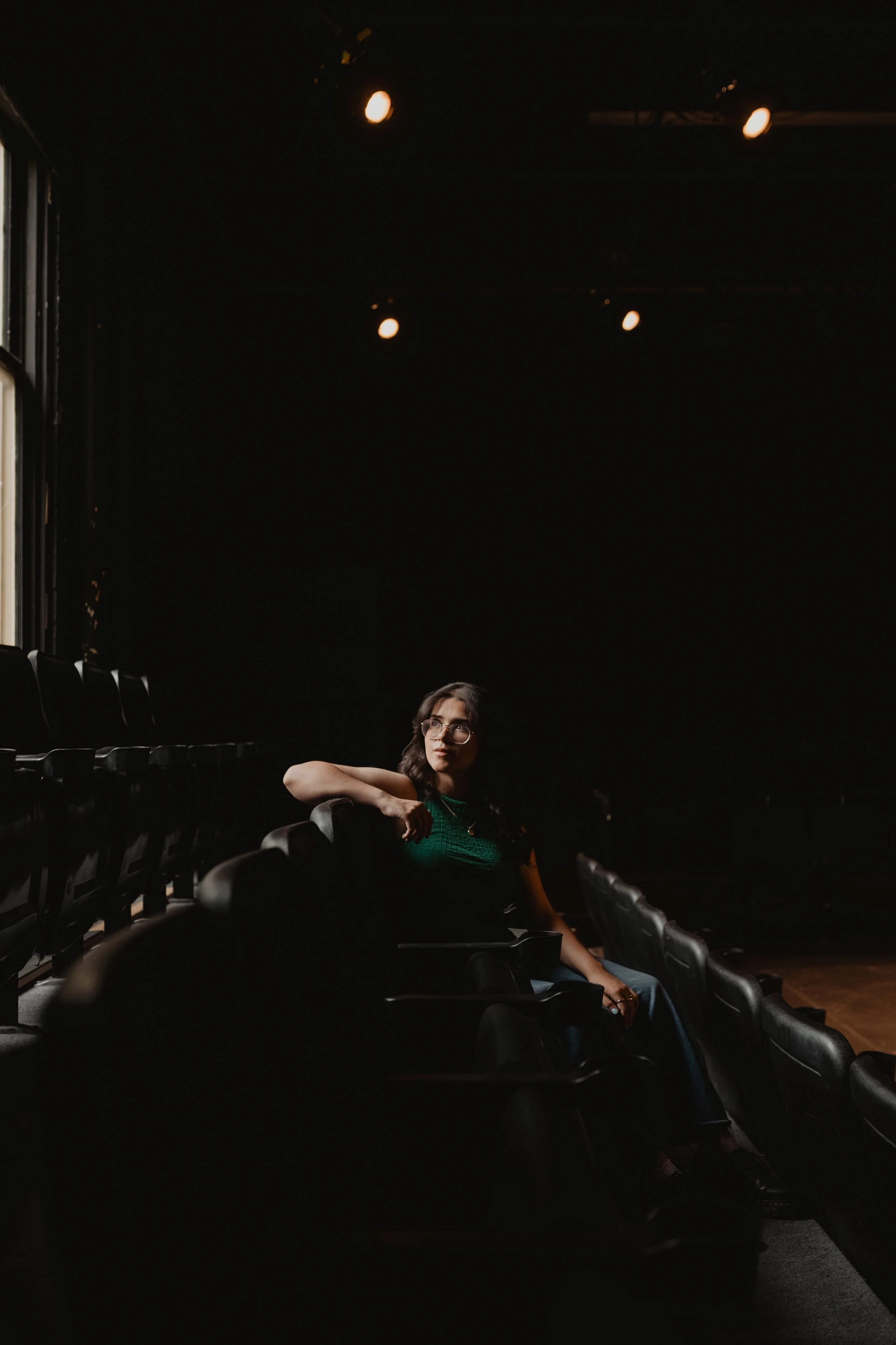 A woman with glasses and dark hair sits alone in an empty dark auditorium, wearing a green top, with only a few overhead lights illuminating the space.