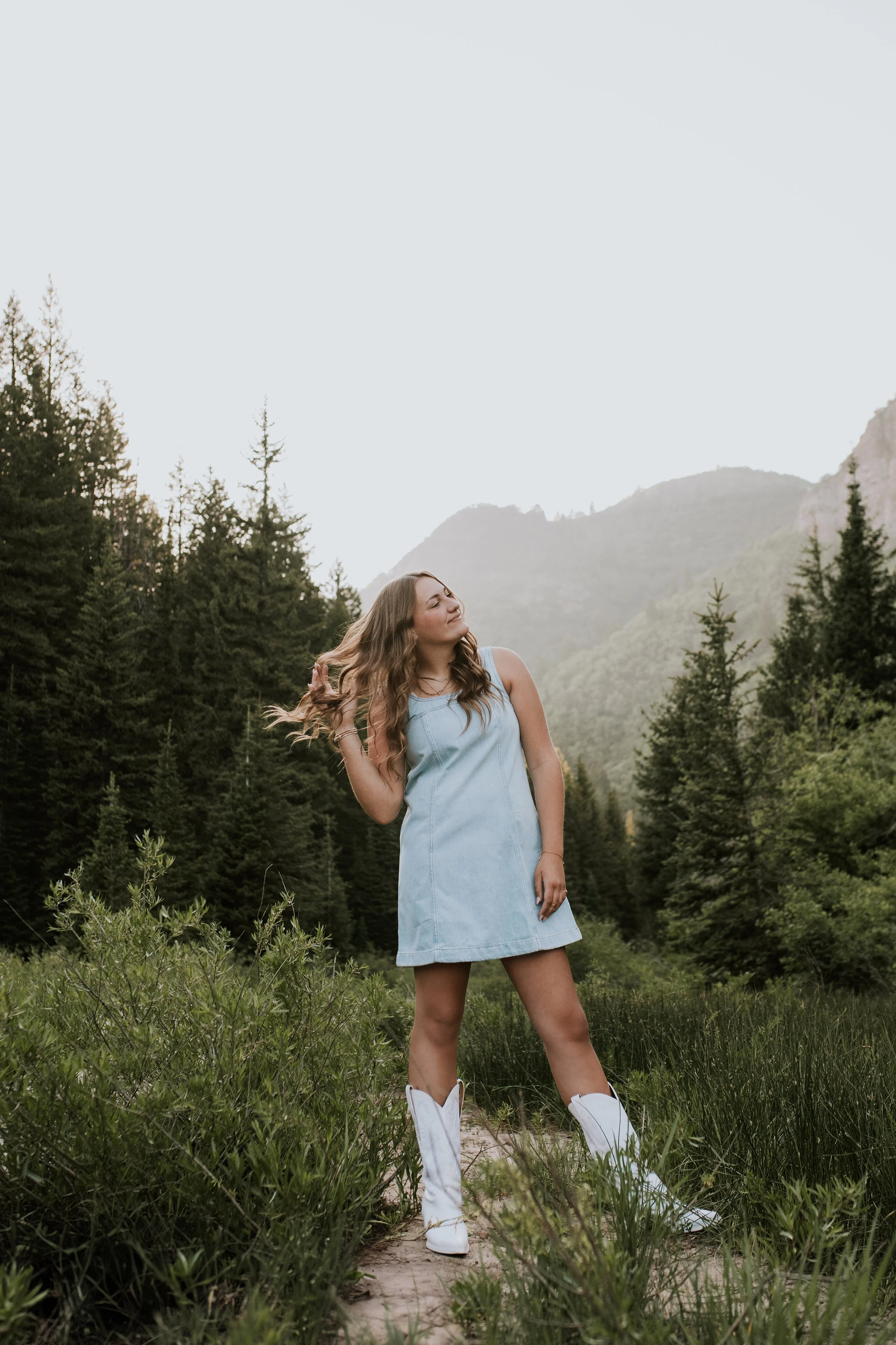 A woman in a light blue dress and white cowboy boots stands outdoors in a lush green forested area with mountains in the background, smiling and touching her hair.