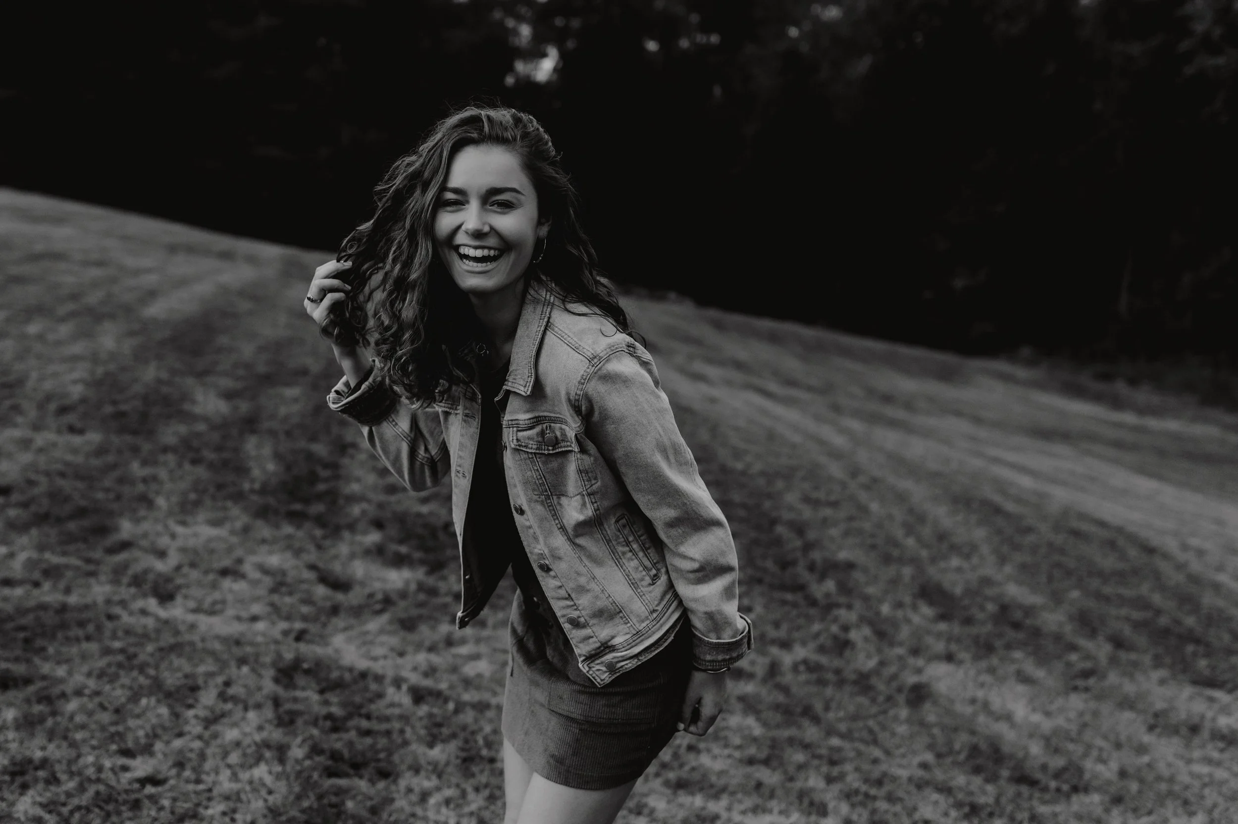 Black and white photo of a smiling young woman with curly hair, wearing a denim jacket and a skirt, standing outdoors on grass.
