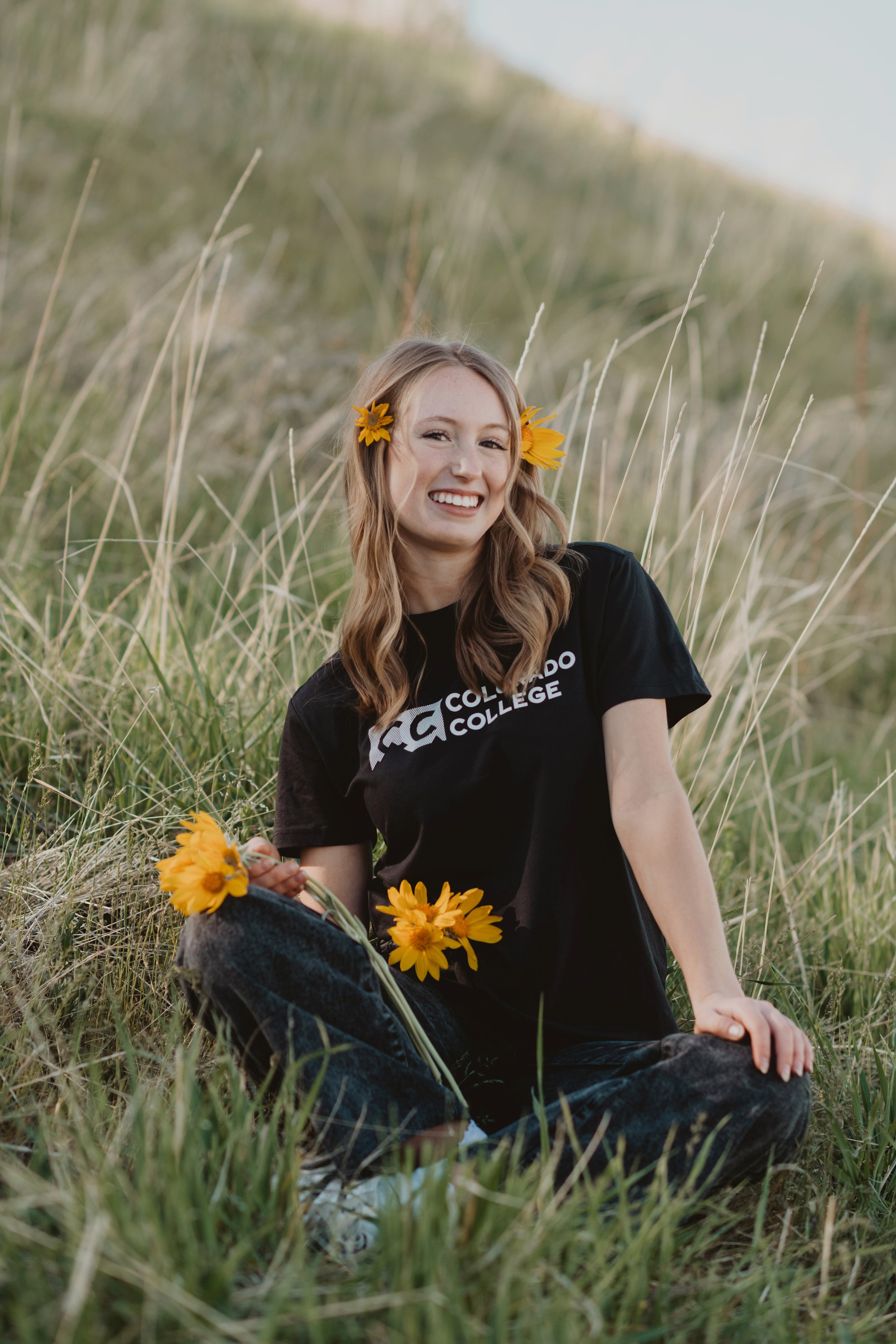 A young woman with long wavy hair, wearing a black ACO college t-shirt, sitting in tall grass and holding yellow flowers, with two yellow flowers tucked behind her ears, smiling at the camera, in an outdoor setting.