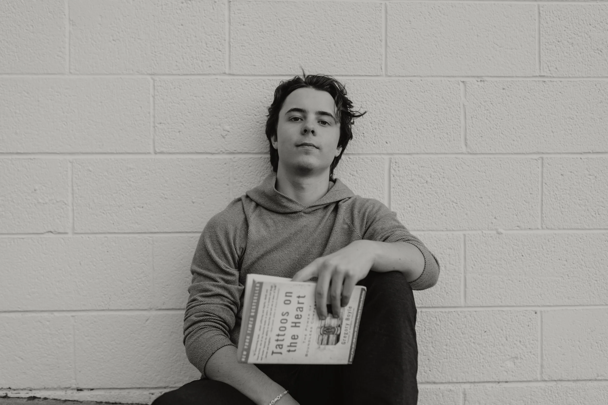 A young person with dark, wavy hair sitting against a light-colored cinder block wall, holding a book titled 'Tattoos on the Heart' by Gregory Boyle in their lap.