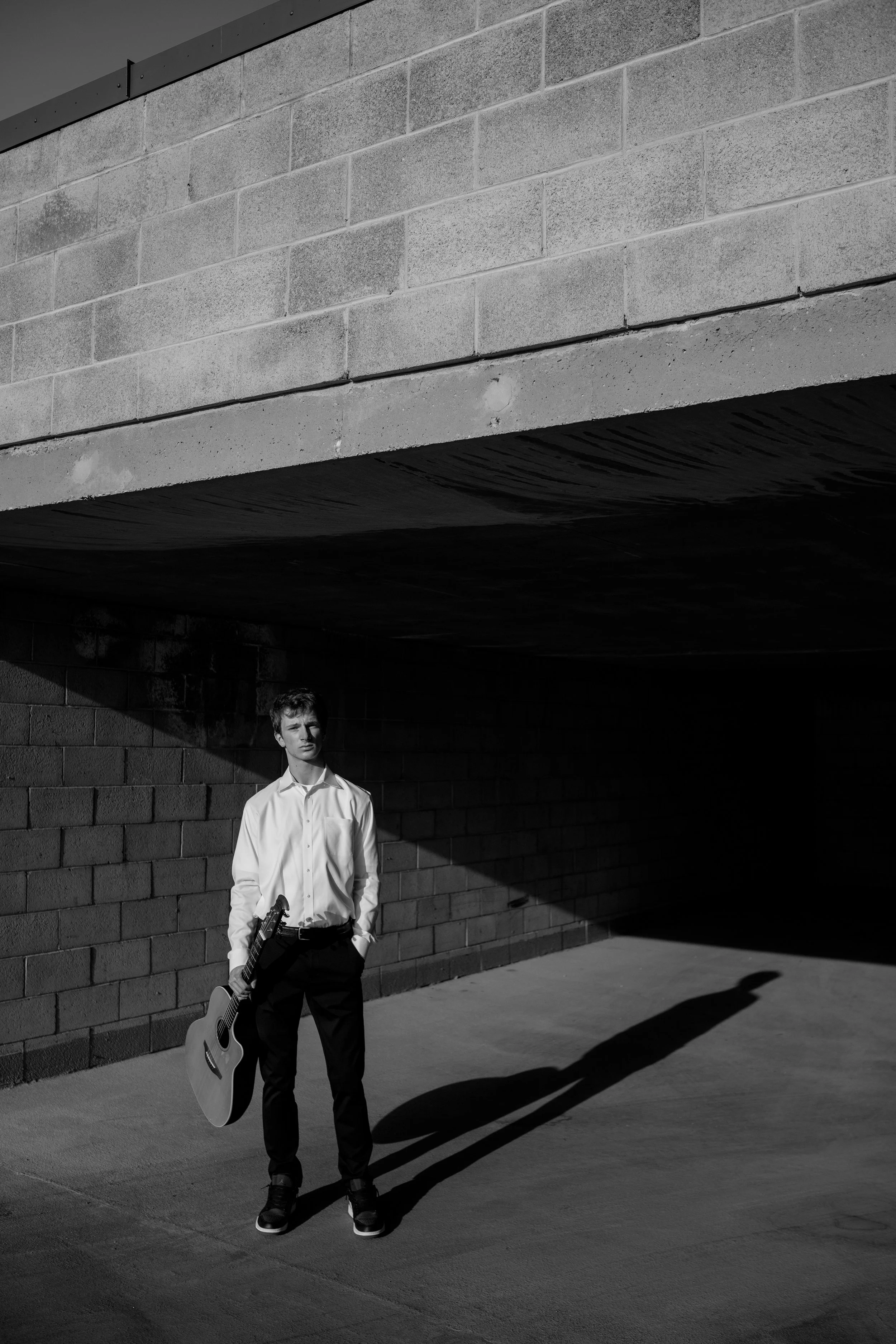 A young man in a white shirt and dark pants holding a guitar, standing against a brick wall with shadows cast on the ground and wall.