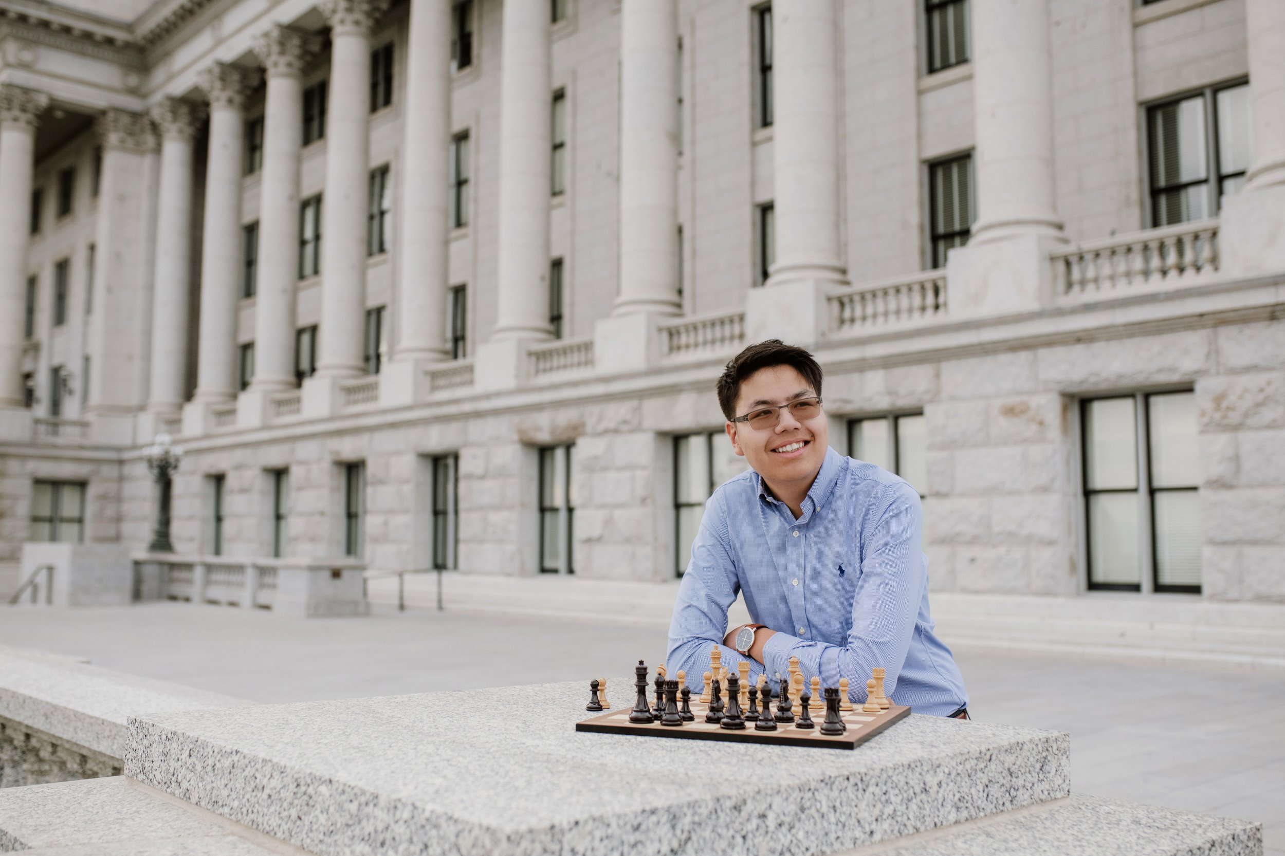 A young man wearing glasses and a blue shirt playing chess outdoors in front of a historic building with large columns.