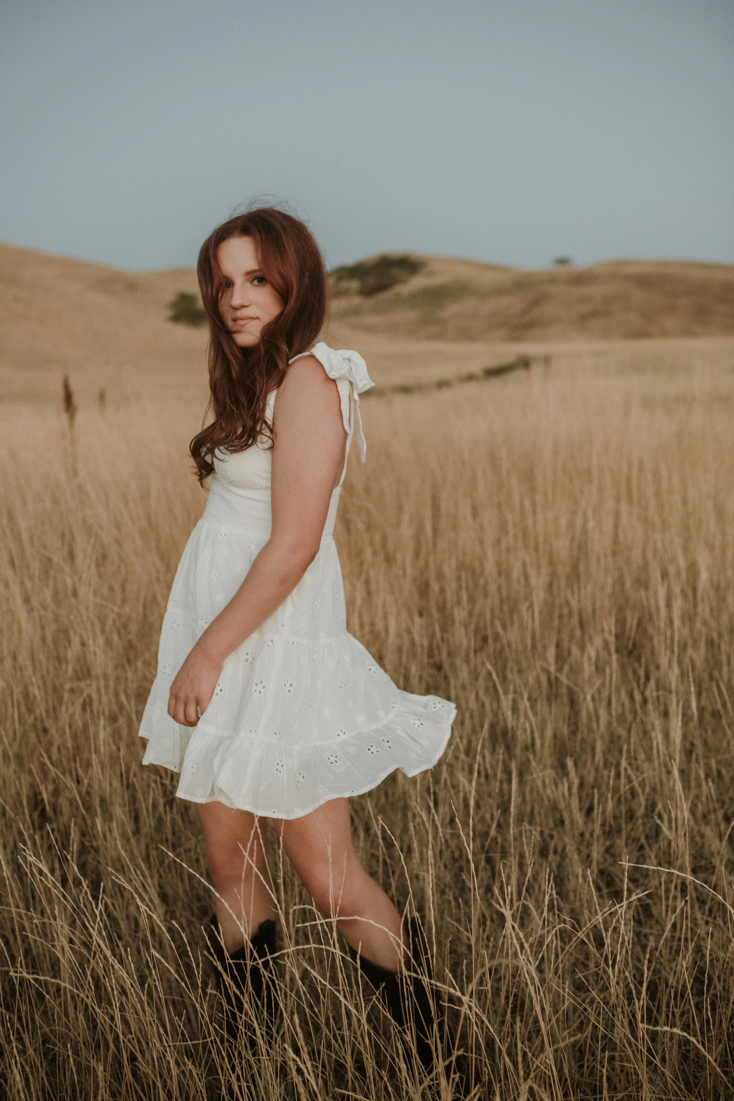 A young woman with red hair wearing a white dress stands in a golden field with rolling hills in the background during dusk or dawn.