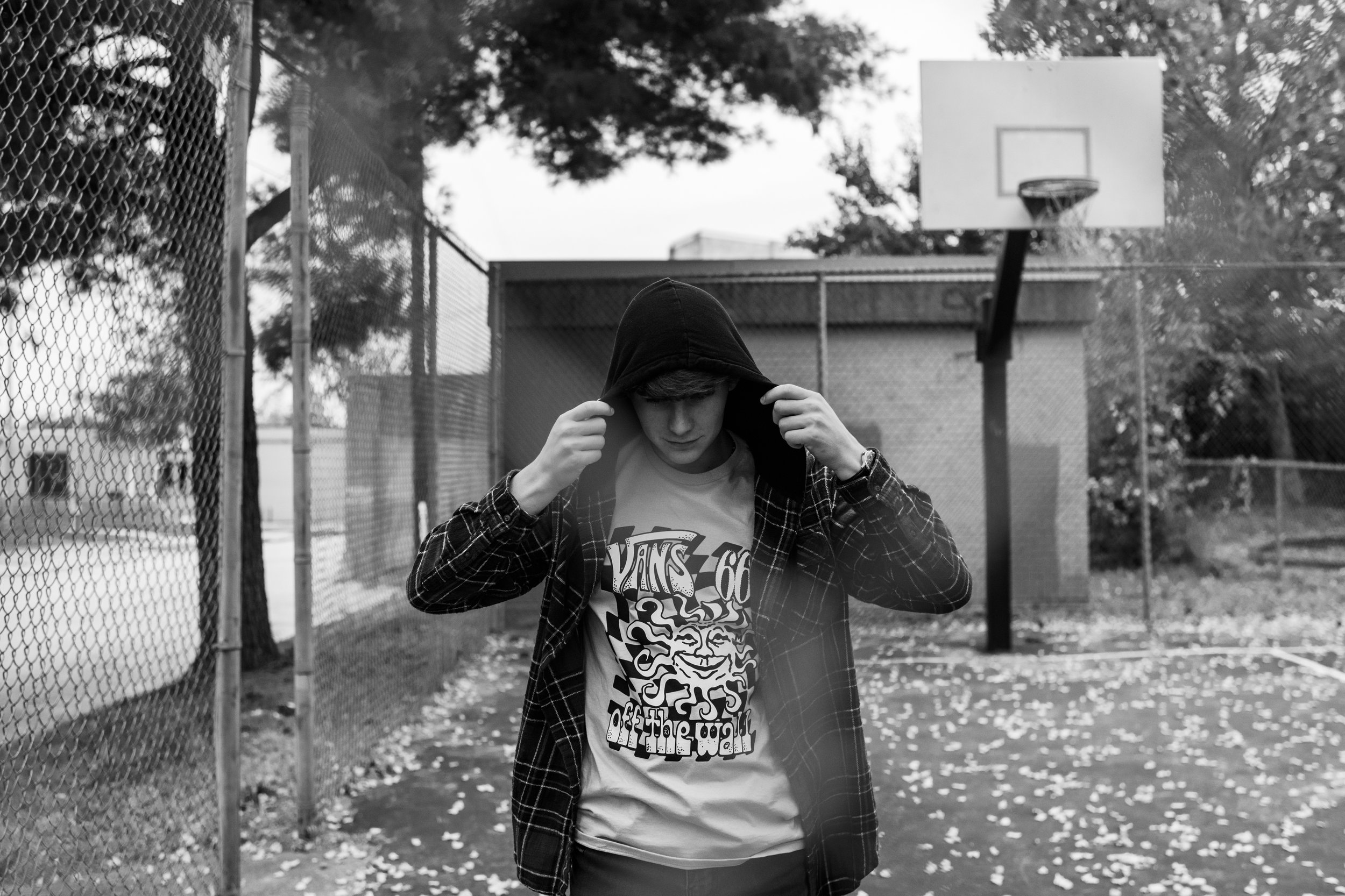 A young person with a hoodie, looking down, walking in an outdoor basketball court with a fence on one side and trees in the background, black and white photo.