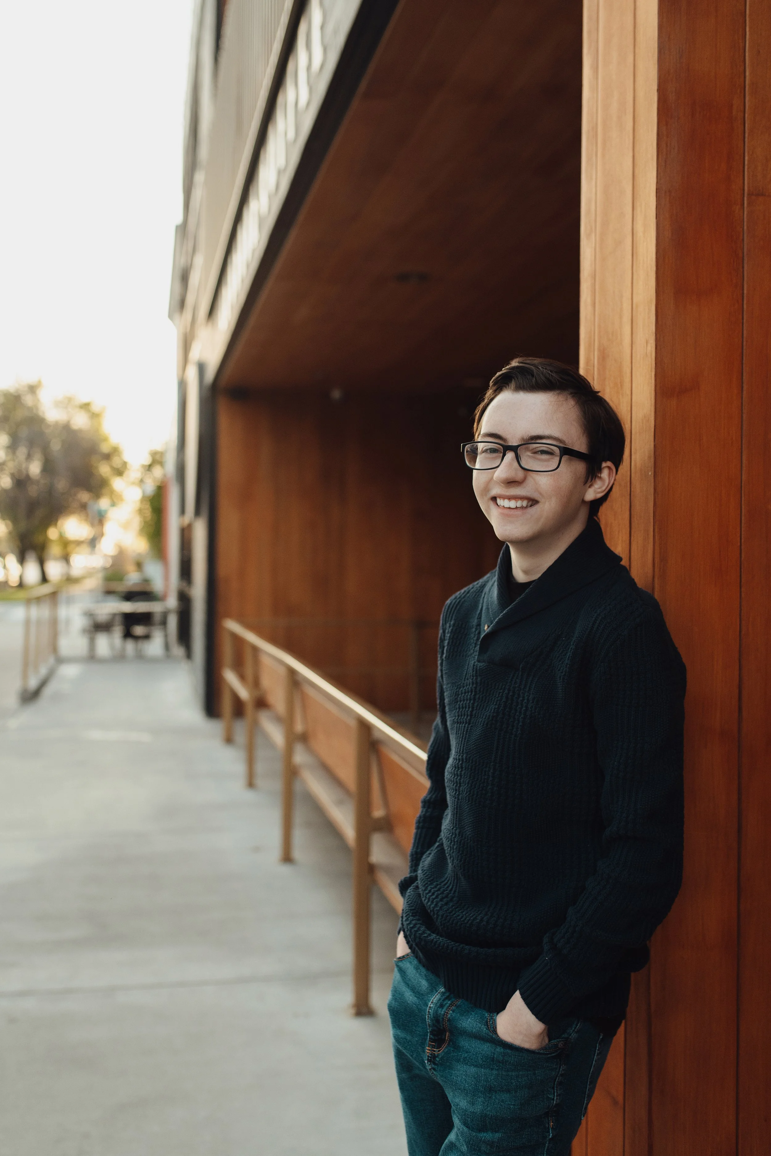 A young man with glasses smiling, leaning against a wooden wall outdoors.