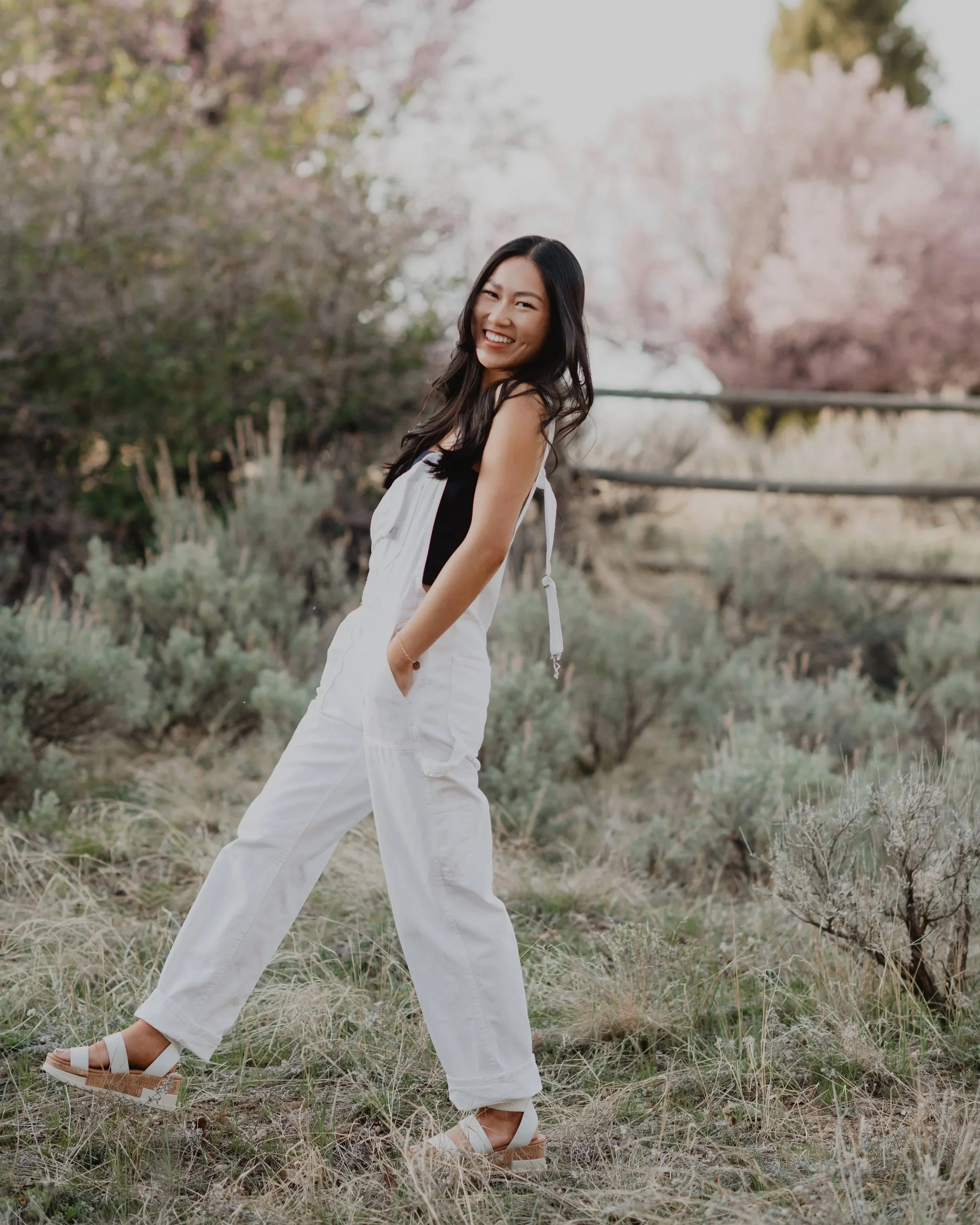 A young woman smiling and posing outdoors in a field with desert plants and trees in the background, wearing white wide-leg pants, a black and white top, and platform sandals.