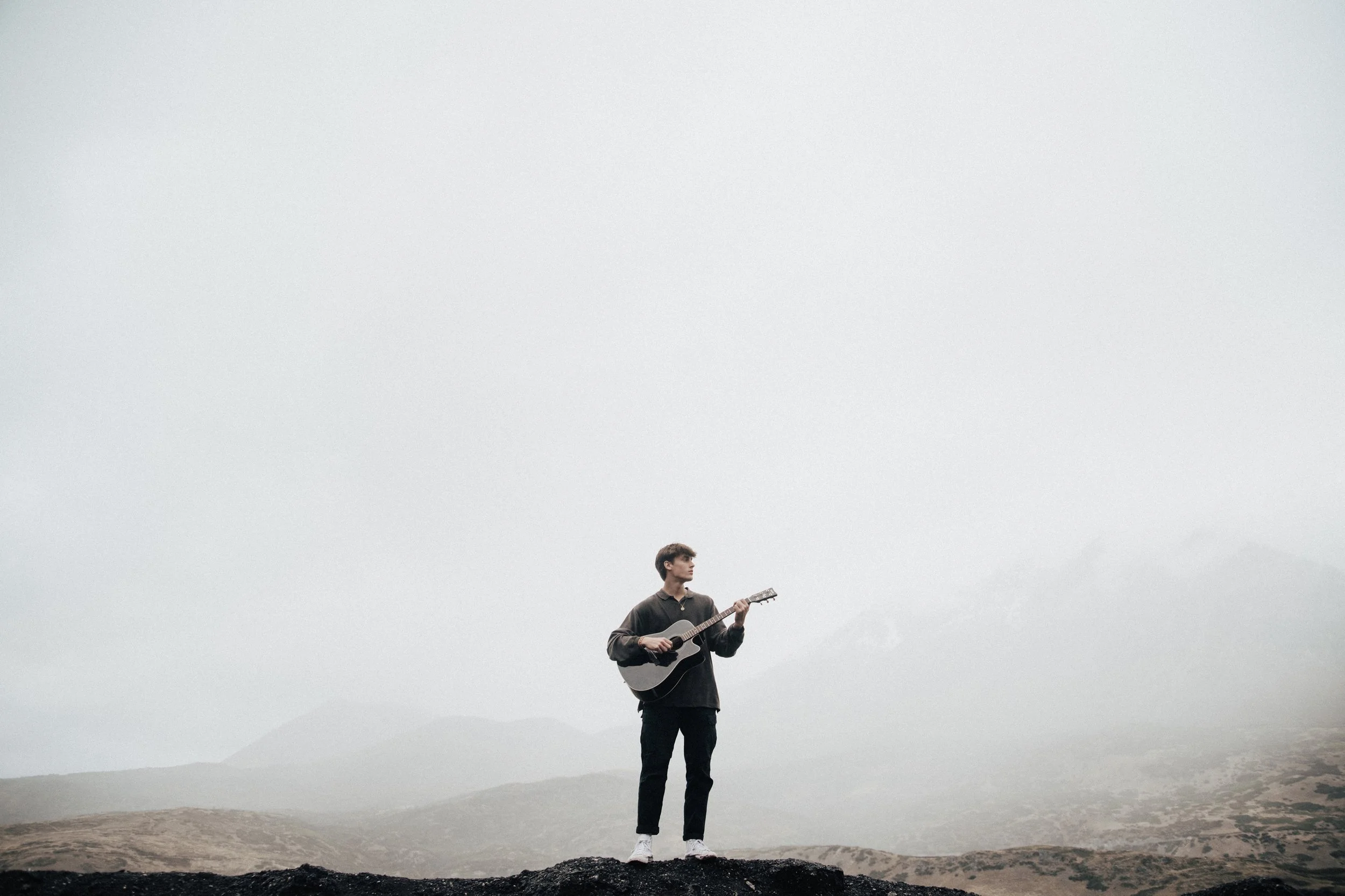 A young man standing outdoors on a rocky terrain, playing an acoustic guitar. The background features a foggy landscape with mountains and a cloudy sky.