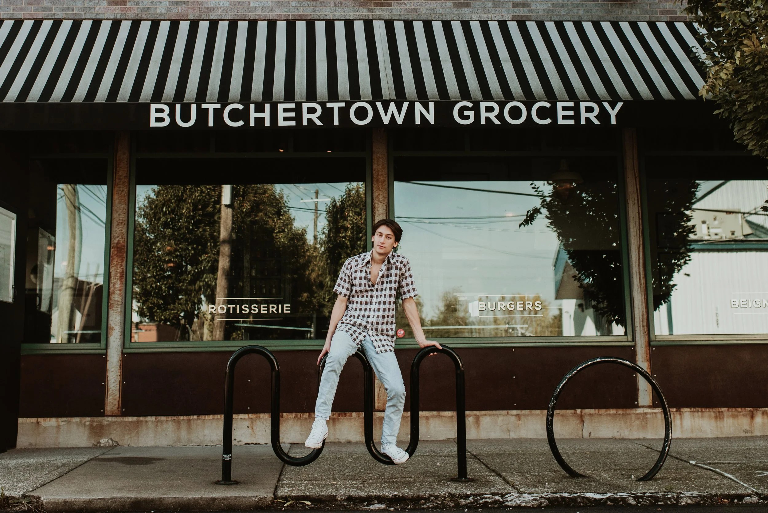 A young person with short dark hair is standing outside a grocery store called 'Butchertown Grocery.' They are wearing a patterned shirt and light jeans, leaning on a bike rack with one hand.