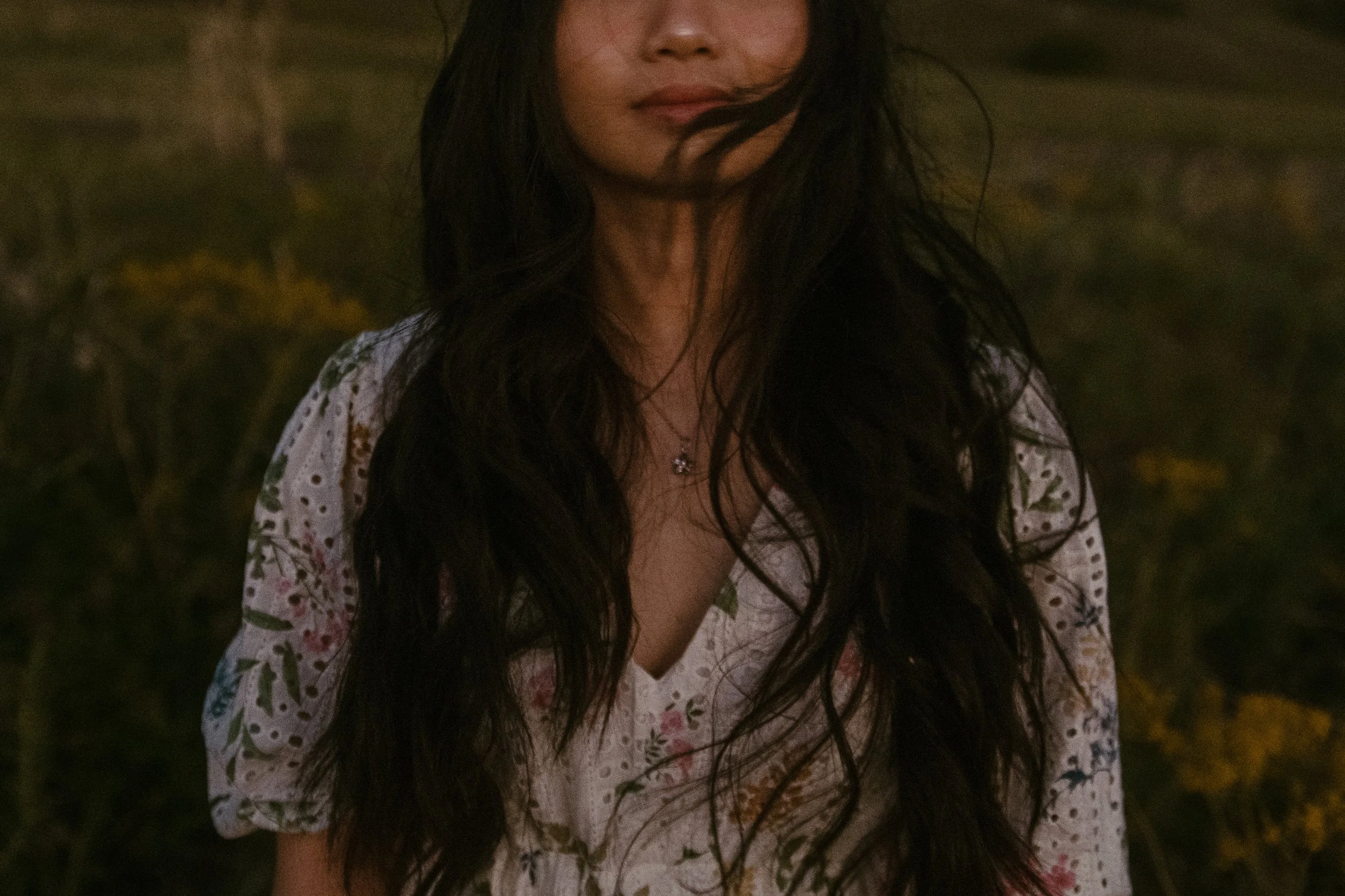 Close-up of a woman with long dark curly hair, wearing a white floral dress with a neckline, and a small necklace, standing outdoors at dusk or evening.