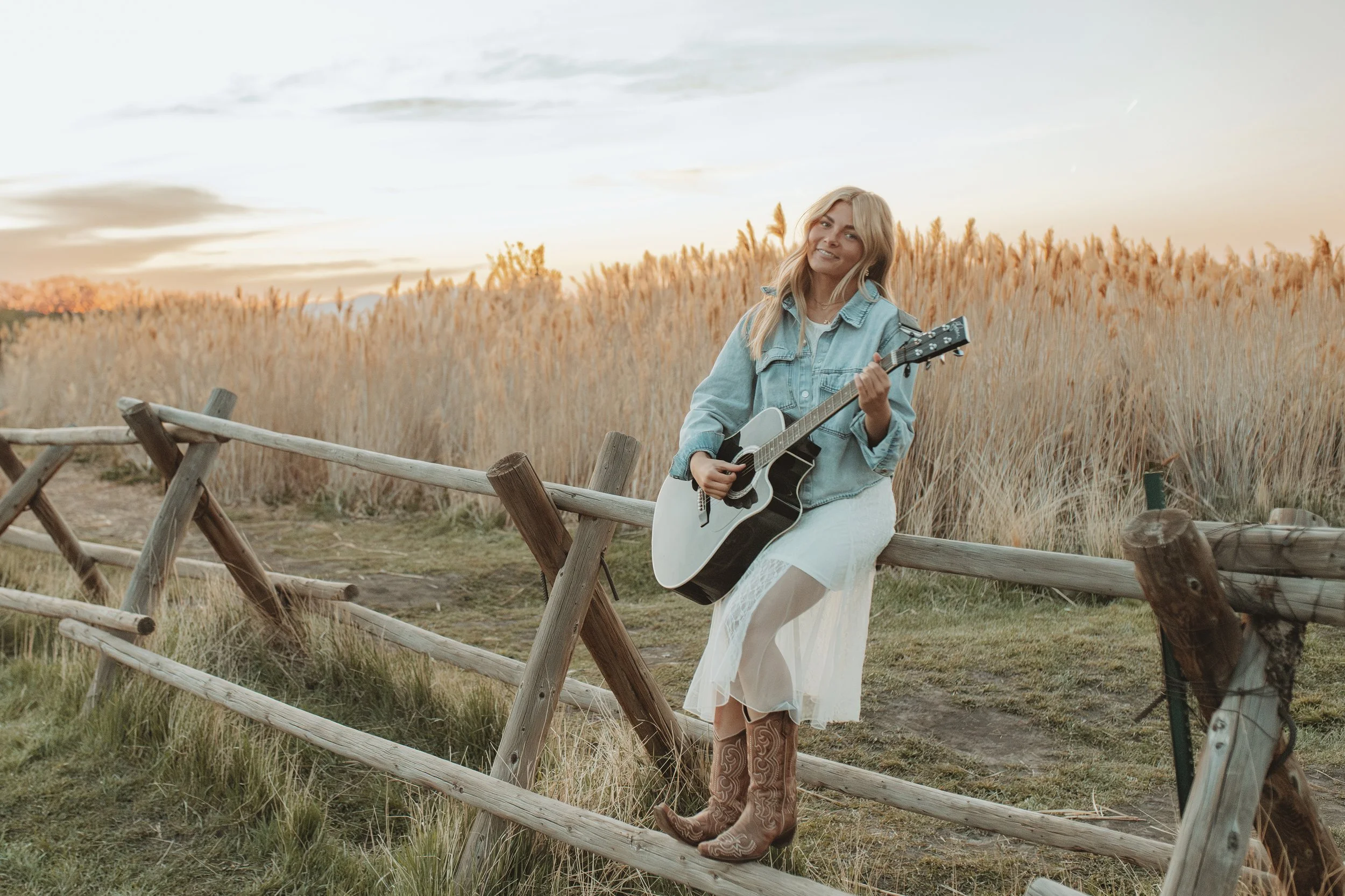 A young woman with long blonde hair wearing a denim jacket, white dress, and cowboy boots, leaning on a rustic wooden fence while playing an acoustic guitar in a field of tall golden grass during sunset.