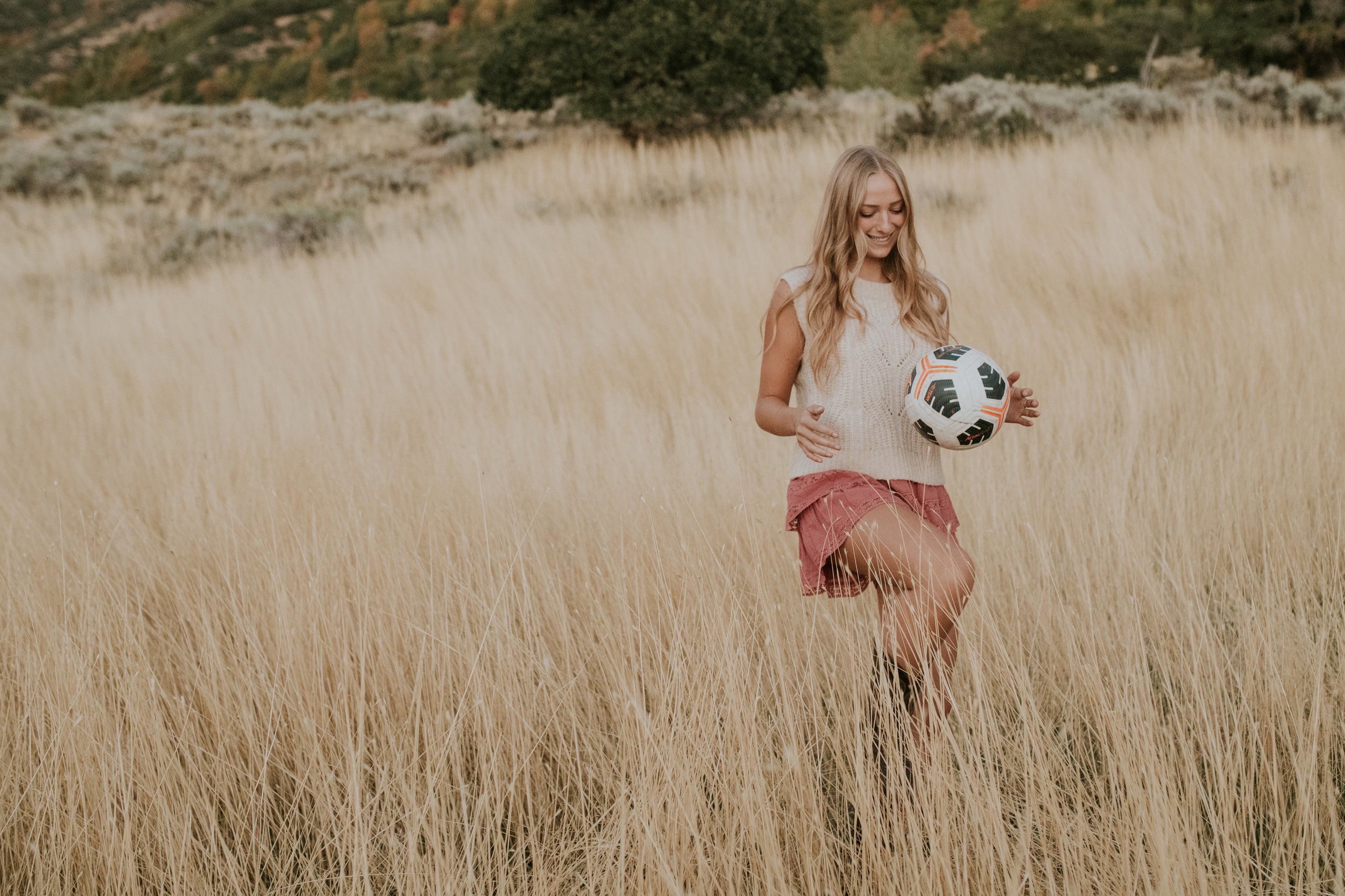 A young woman standing in a field of tall, dry grass, holding a soccer ball, smiling and looking down.
