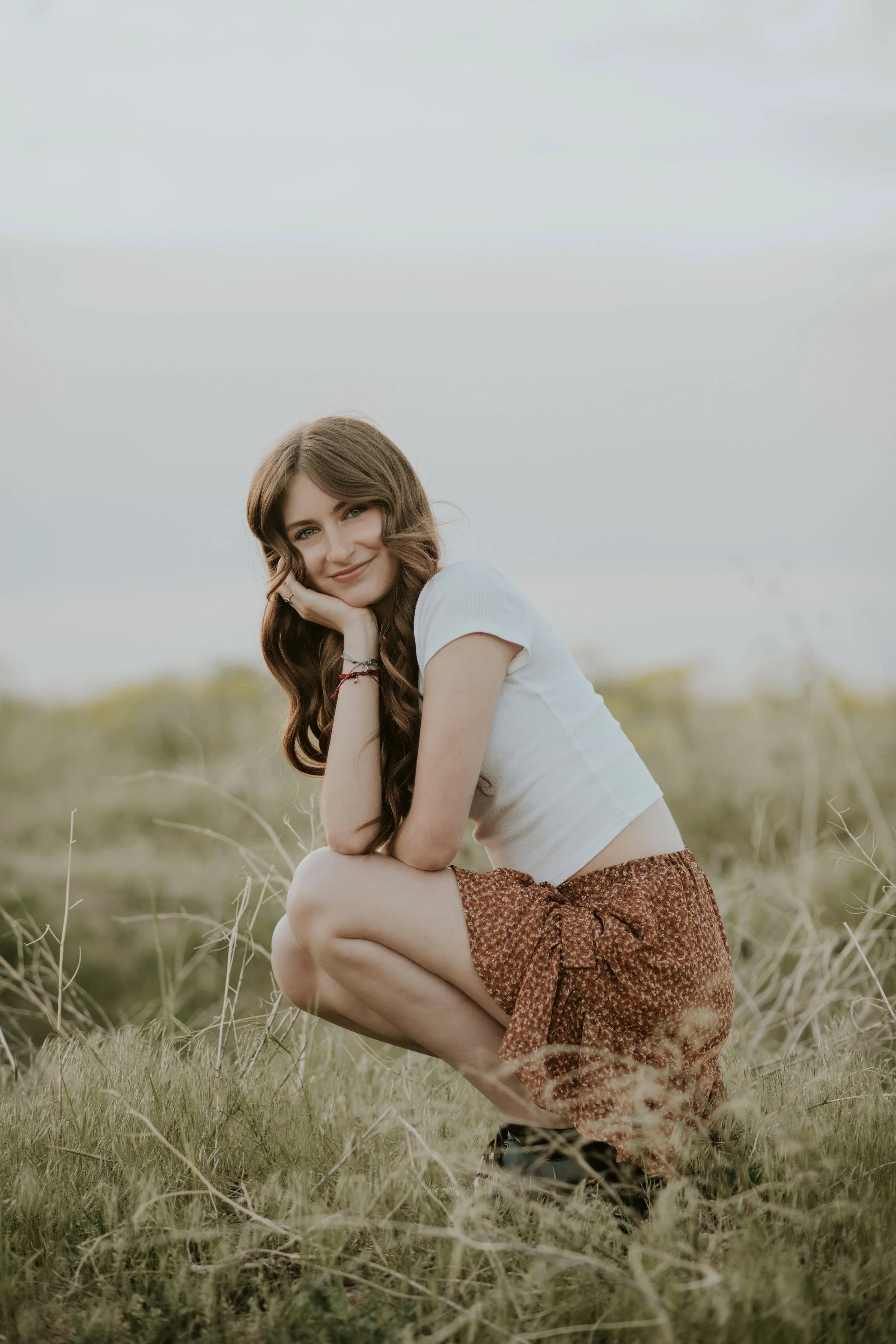 A young woman with long, wavy brown hair squatting in a field of grass and dried plants, smiling at the camera with her head resting on her hand. She wears a white t-shirt and a patterned brown skirt.