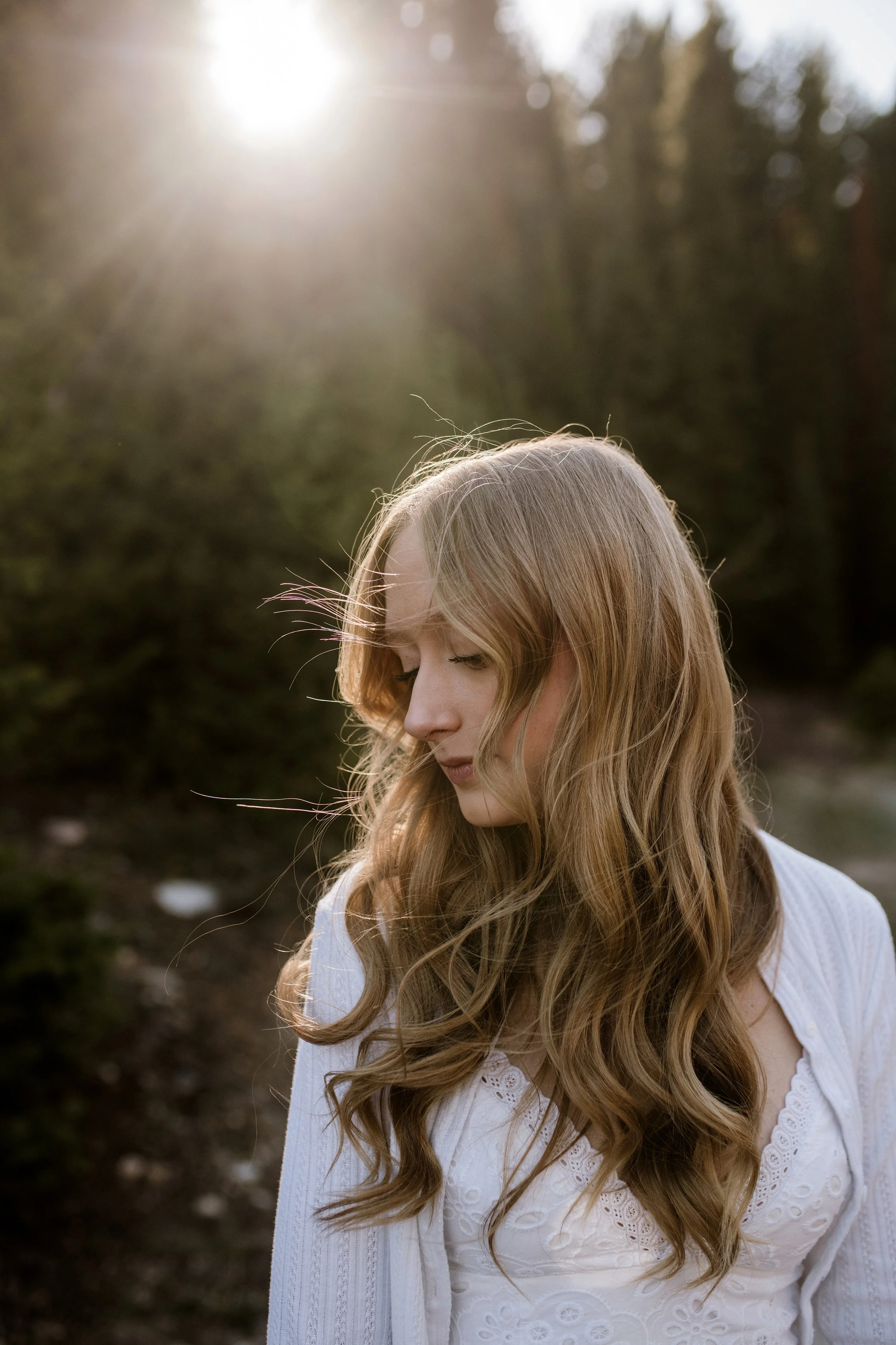 A woman with long, curly blonde hair wearing a white, embroidery dress, standing outdoors with sunlight streaming overhead and trees in the background.