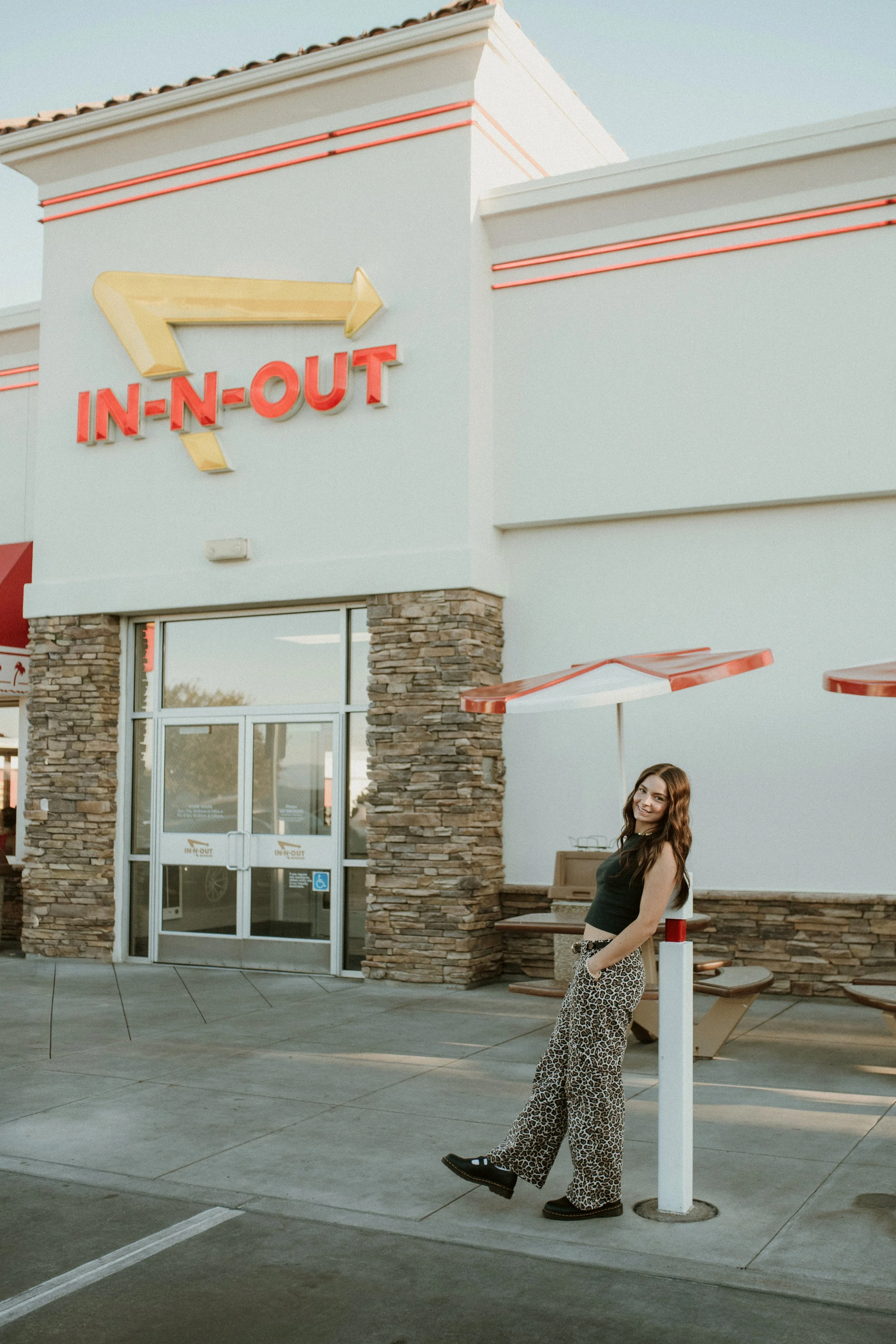 A young woman with long brown hair, wearing leopard print pants and a black top, is smiling and standing outside a fast-food restaurant called In-N-Out Burger. The restaurant has a light-colored exterior with stone accents, and red neon lights on the