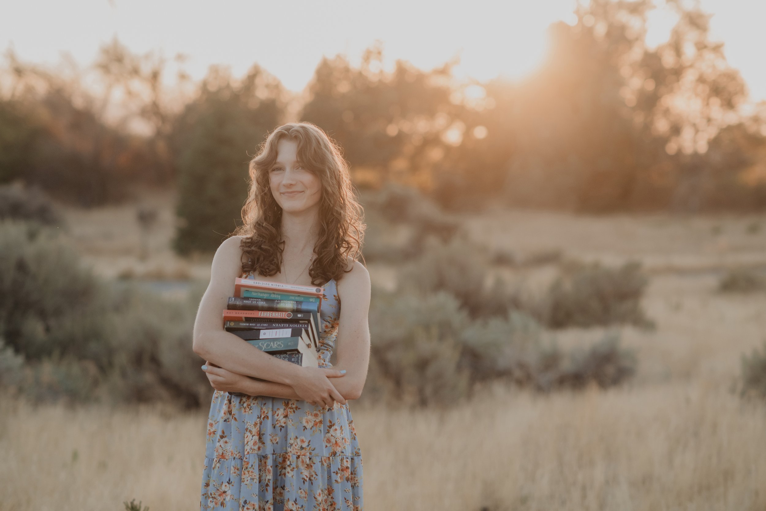Young woman with curly hair holding a stack of books in a field at sunset.