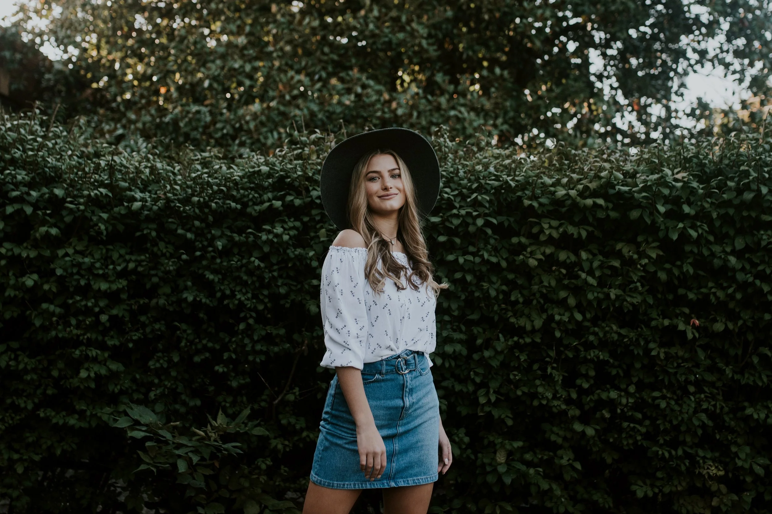 Young woman standing outdoors in front of greenery, wearing a white off-the-shoulder blouse, a black wide-brim hat, and denim skirt.