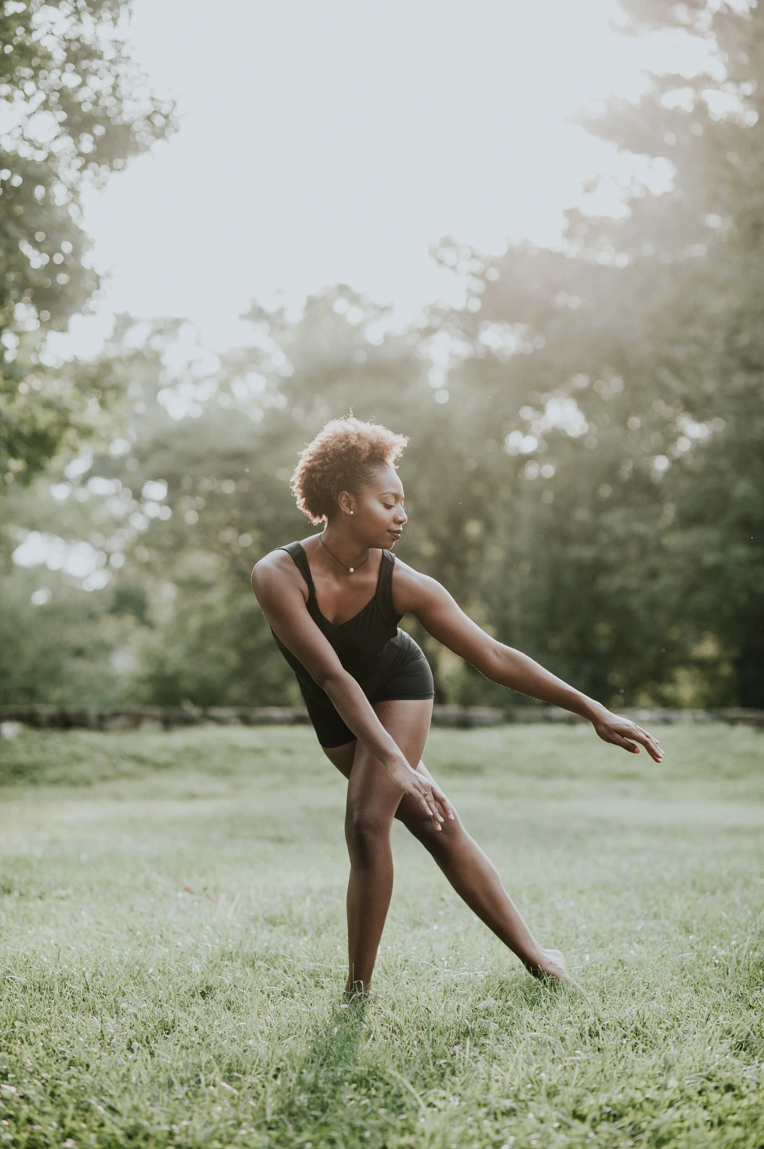 A woman posing in a dance movement outdoors on a grassy field with trees in the background during sunset.