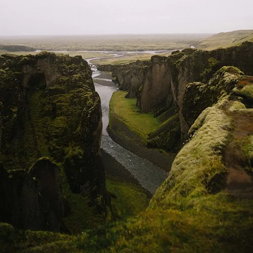 A river flowing through a deep canyon with moss-covered cliffs on either side, overlooking a flat landscape in the distance.