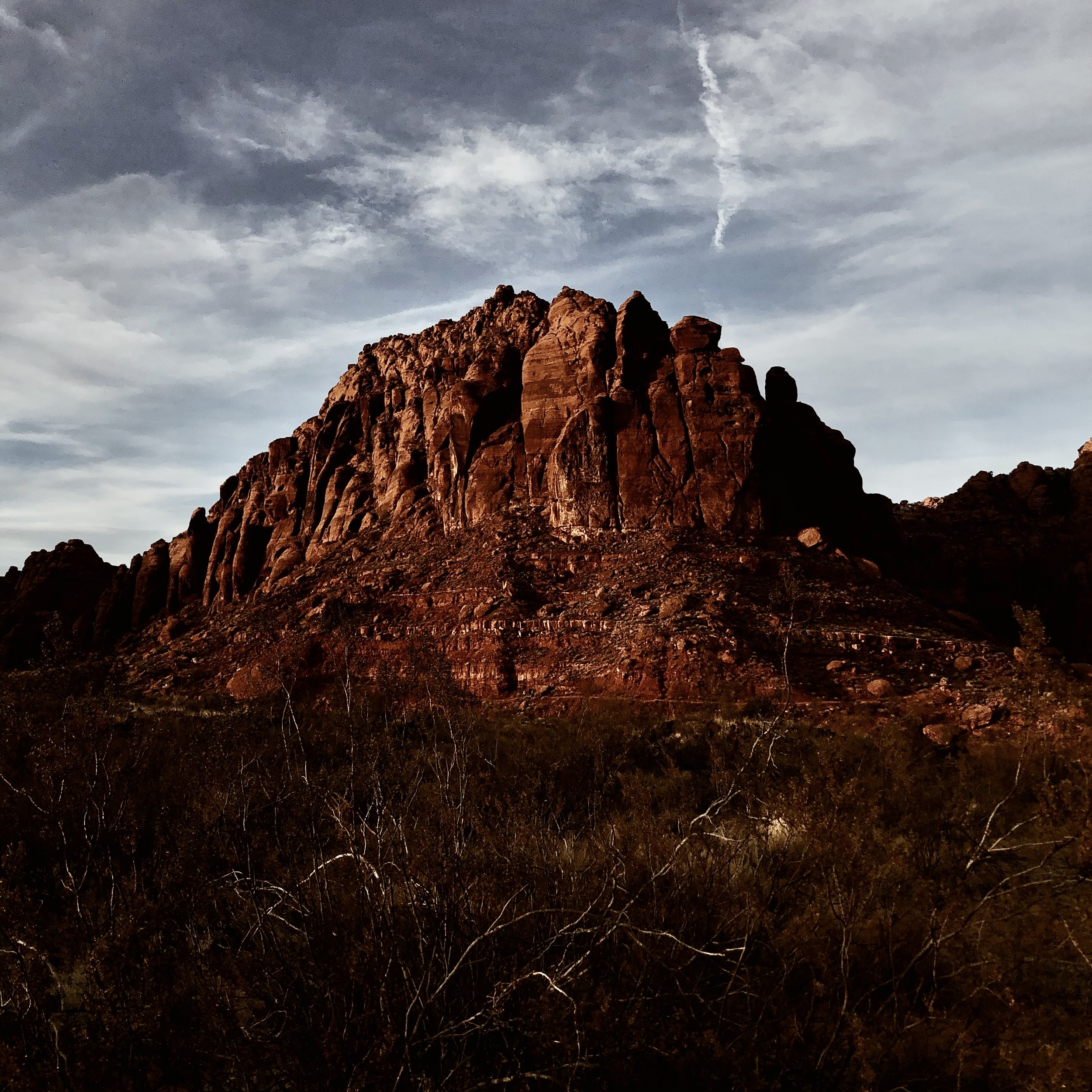 A rugged mountain with reddish-brown rocks against a cloudy sky with streaks of sunlight.