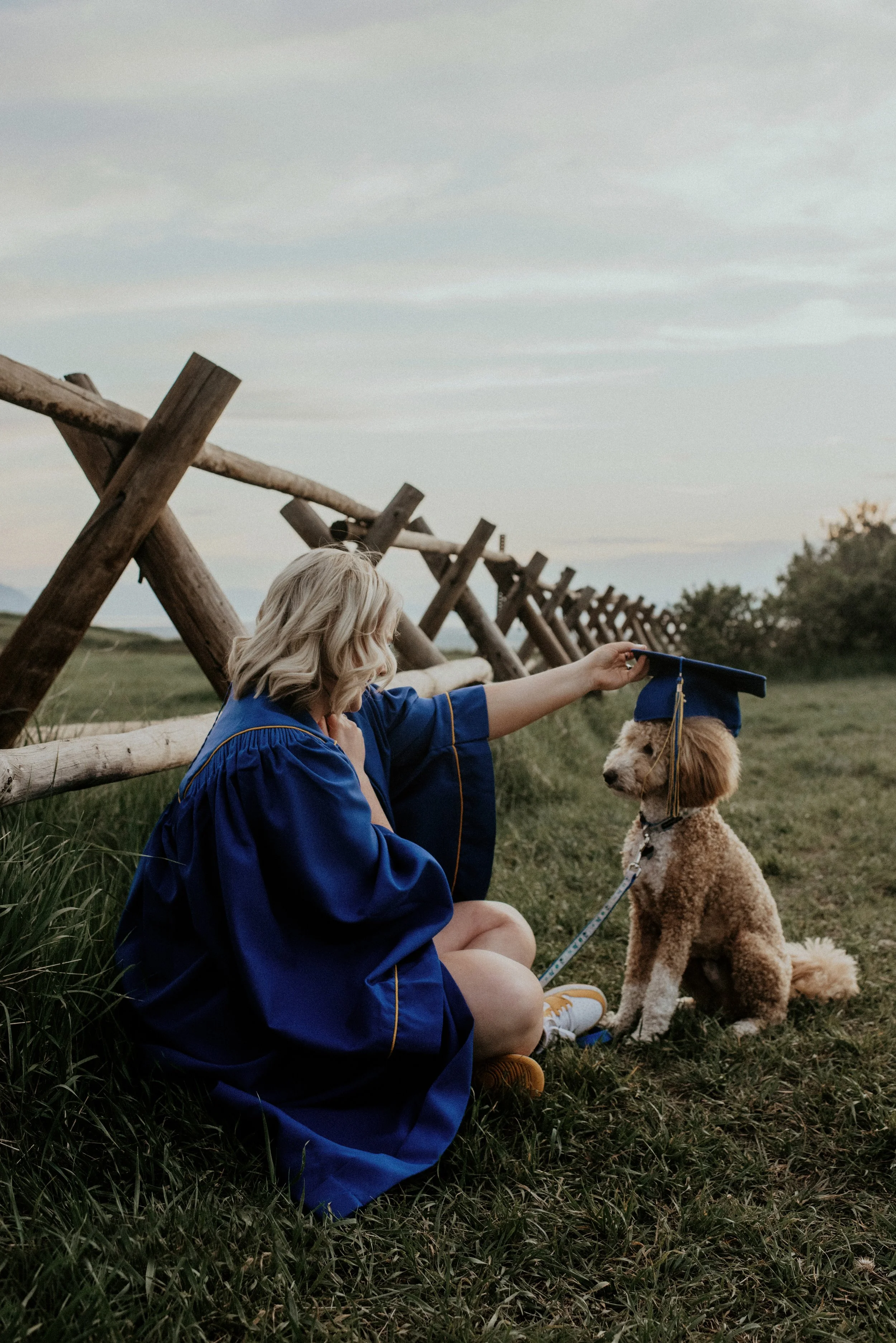 A woman in a blue graduation gown sitting on green grass, with a dog wearing a graduation cap sitting next to her. They are outdoors near a wooden fence with a cloudy sky overhead.