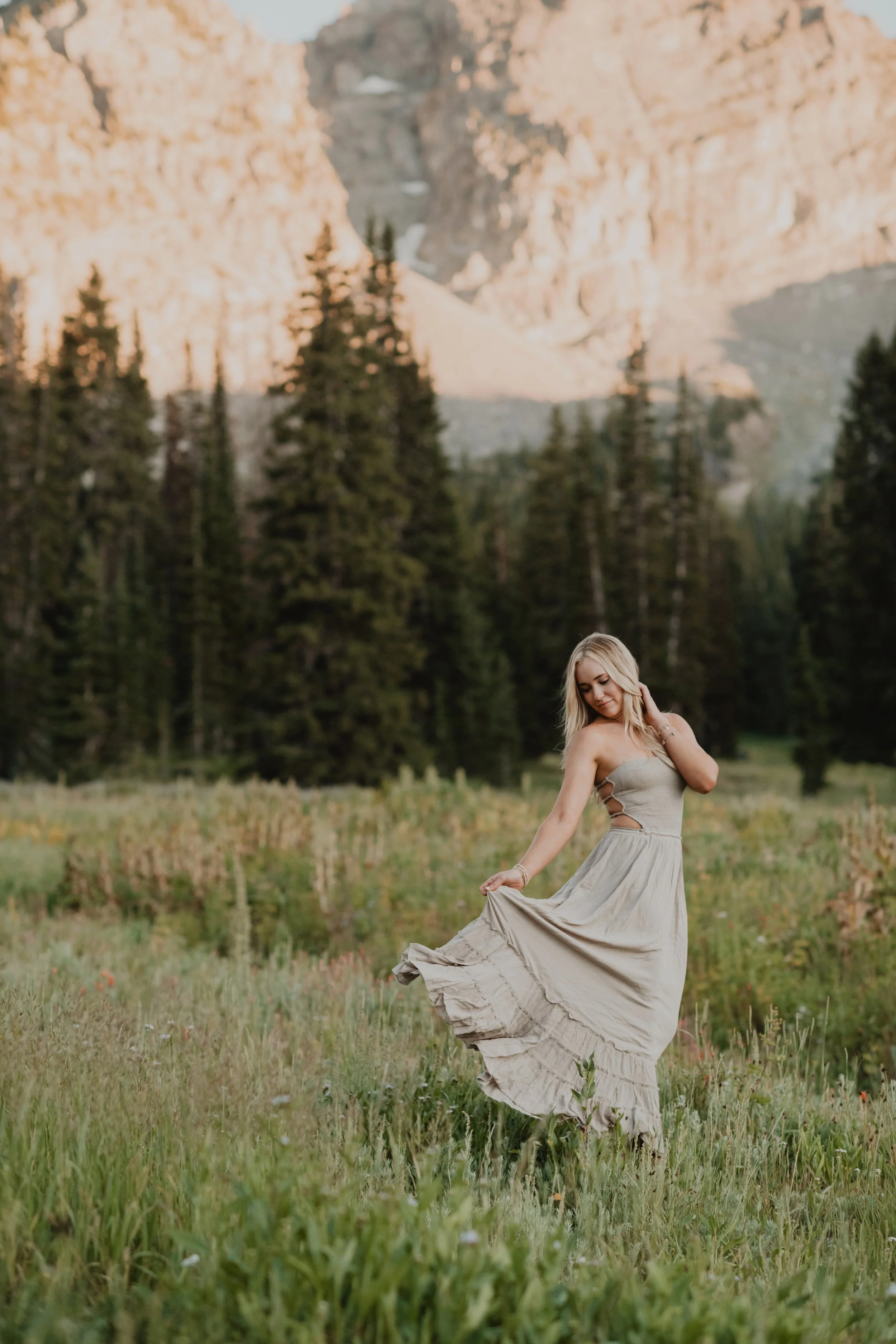 A woman in a beige dress dancing in a grassy field with mountains and evergreen trees in the background.