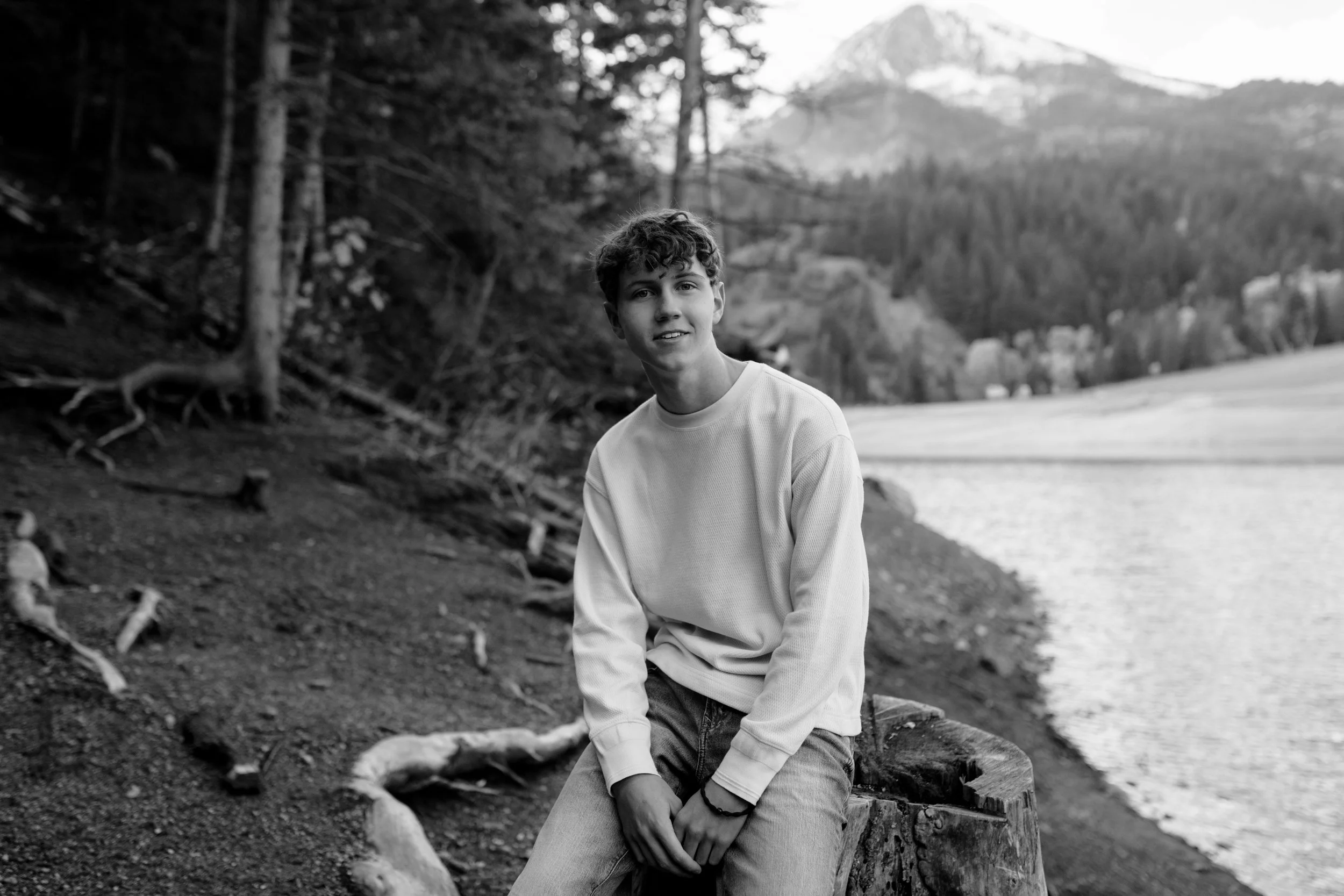 A black and white photo of a young man sitting on a log near a body of water, with trees and mountain in the background.