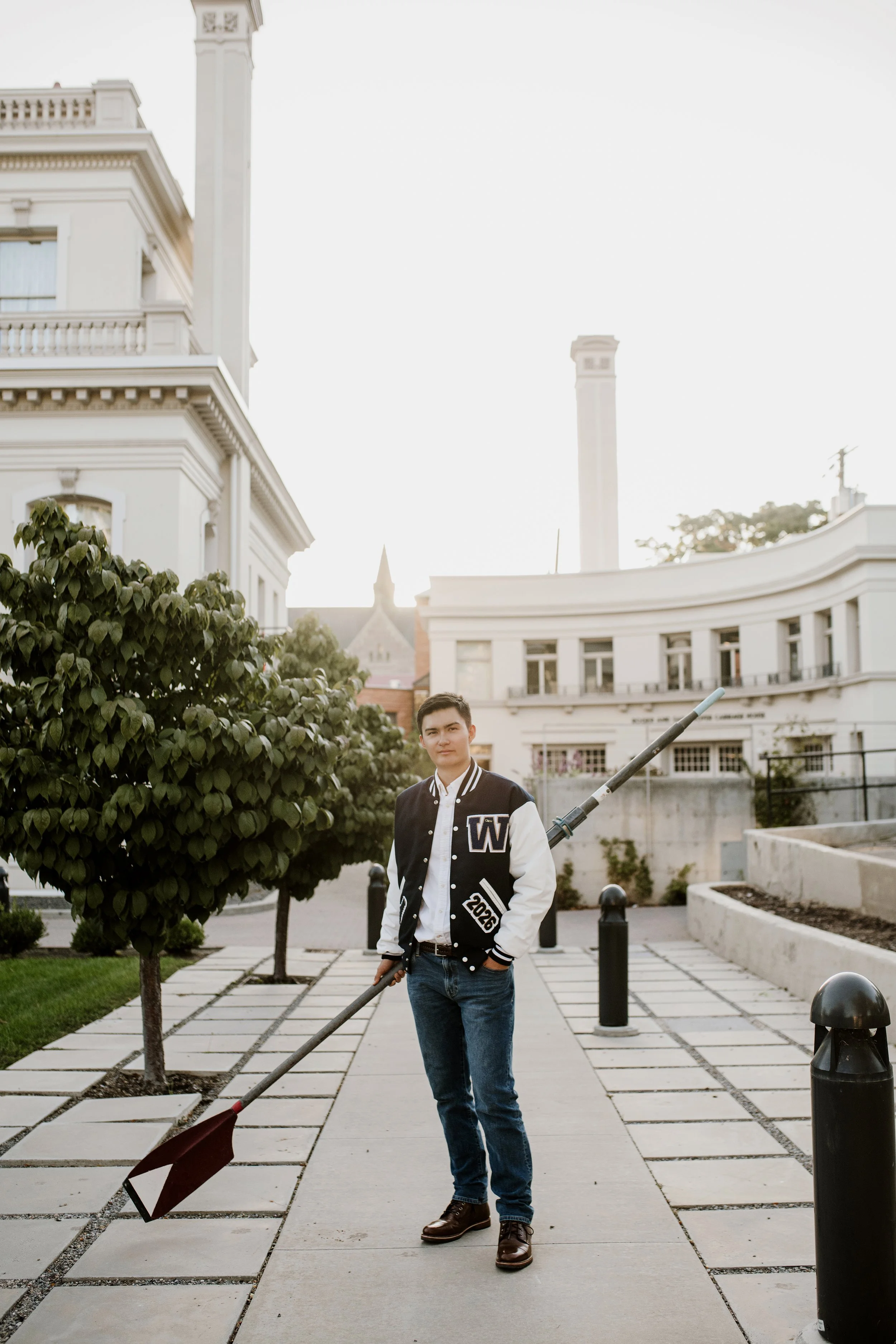 A young man with dark hair in a varsity jacket and jeans standing on a sidewalk holding a gardening tool with a bag attached. There are buildings and greenery around him.