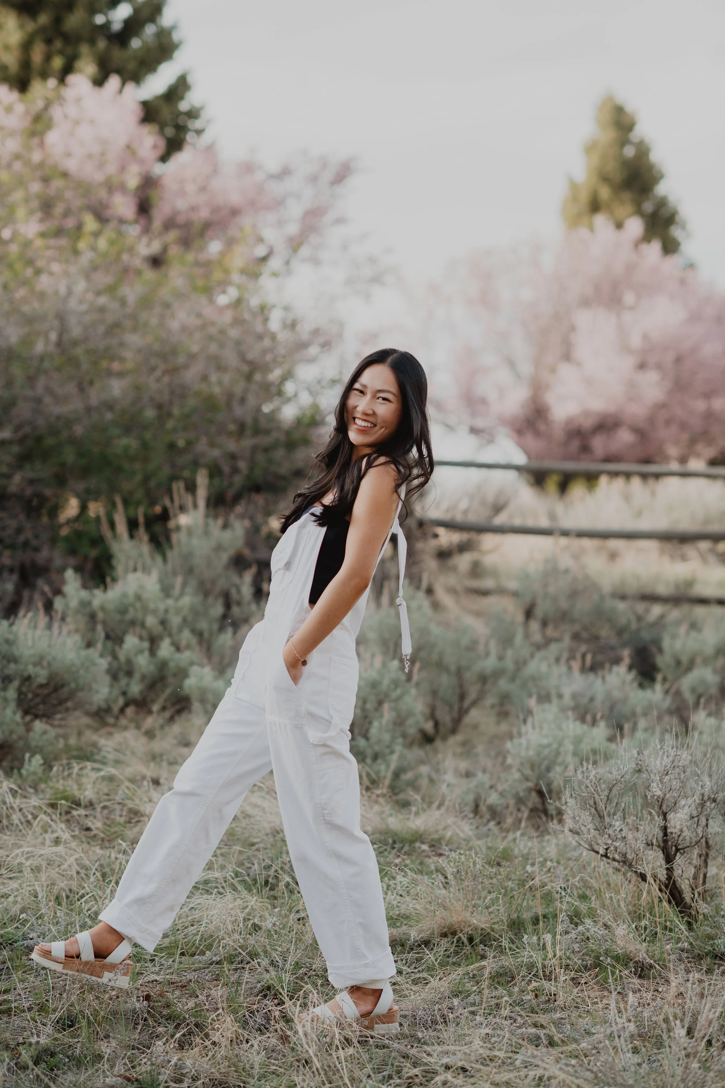 A woman standing in a field with pink flowering trees in the background, smiling and looking at the camera.