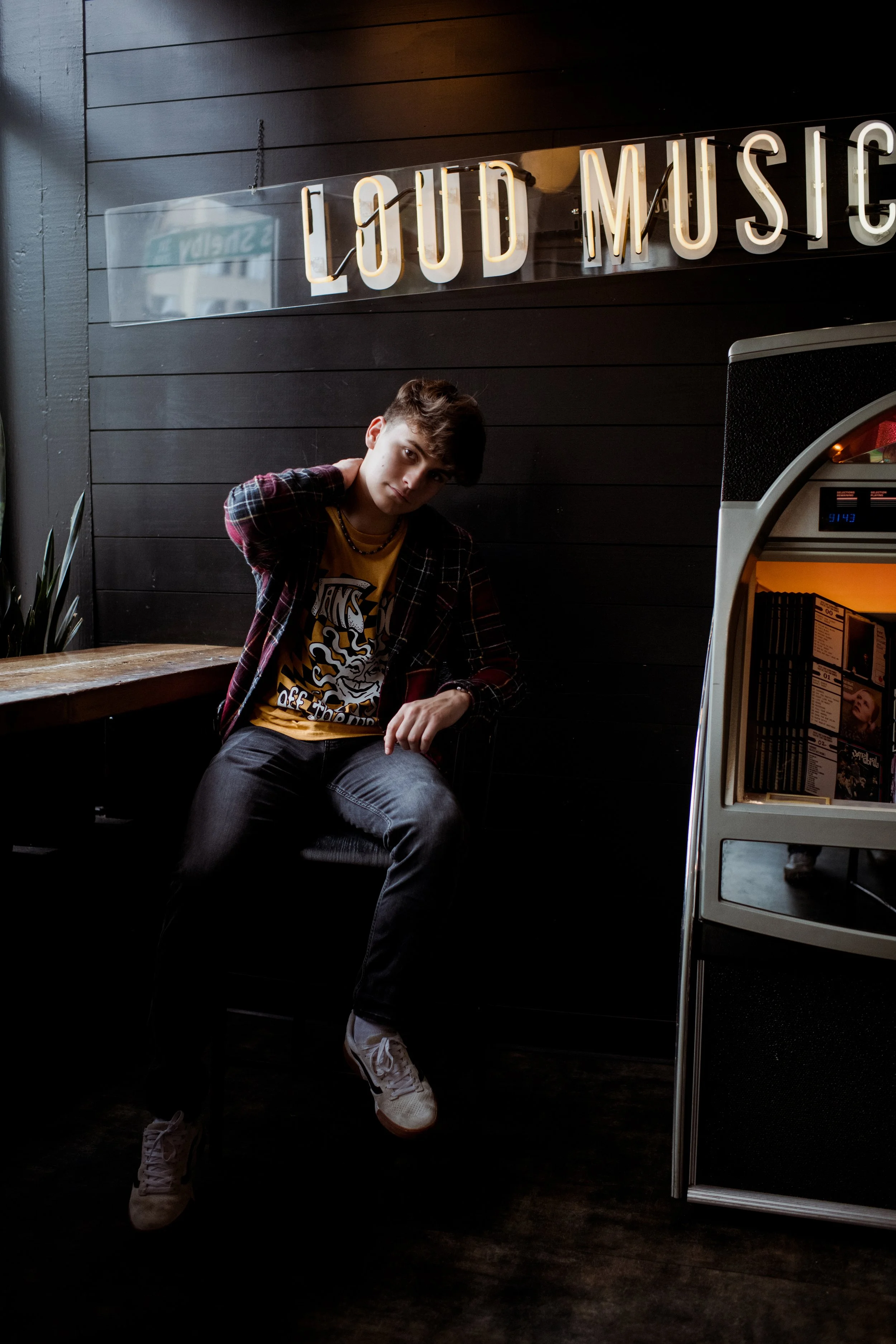A young man with dark hair sitting in a cafe next to a jukebox, under a black sign with illuminated letters that read "LOUD MUSIC." He is wearing a checkered shirt over a graphic t-shirt, dark jeans, and white sneakers, with one hand behind his neck 