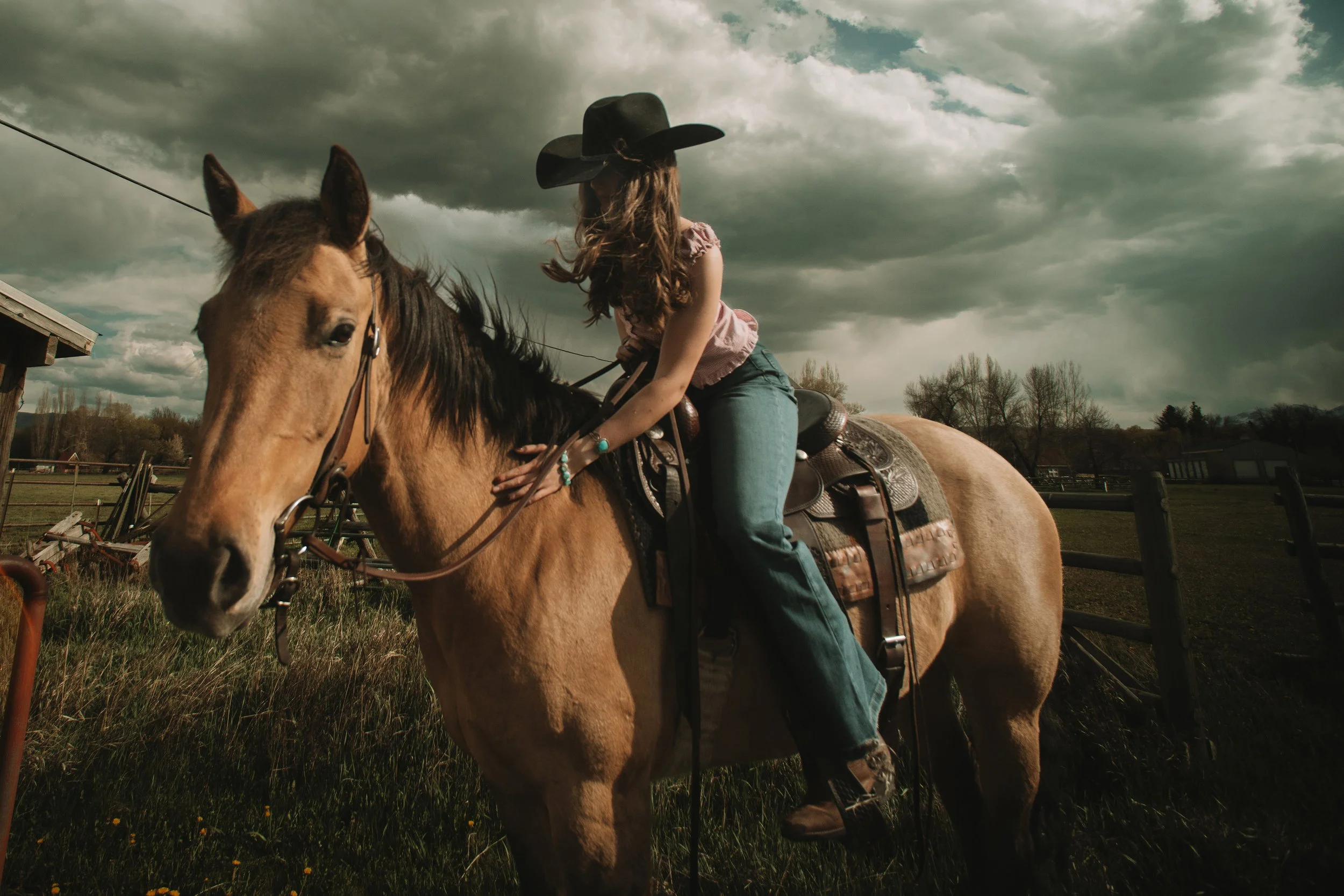 A girl riding a horse outdoors on a cloudy day, wearing a black cowboy hat and casual clothing.