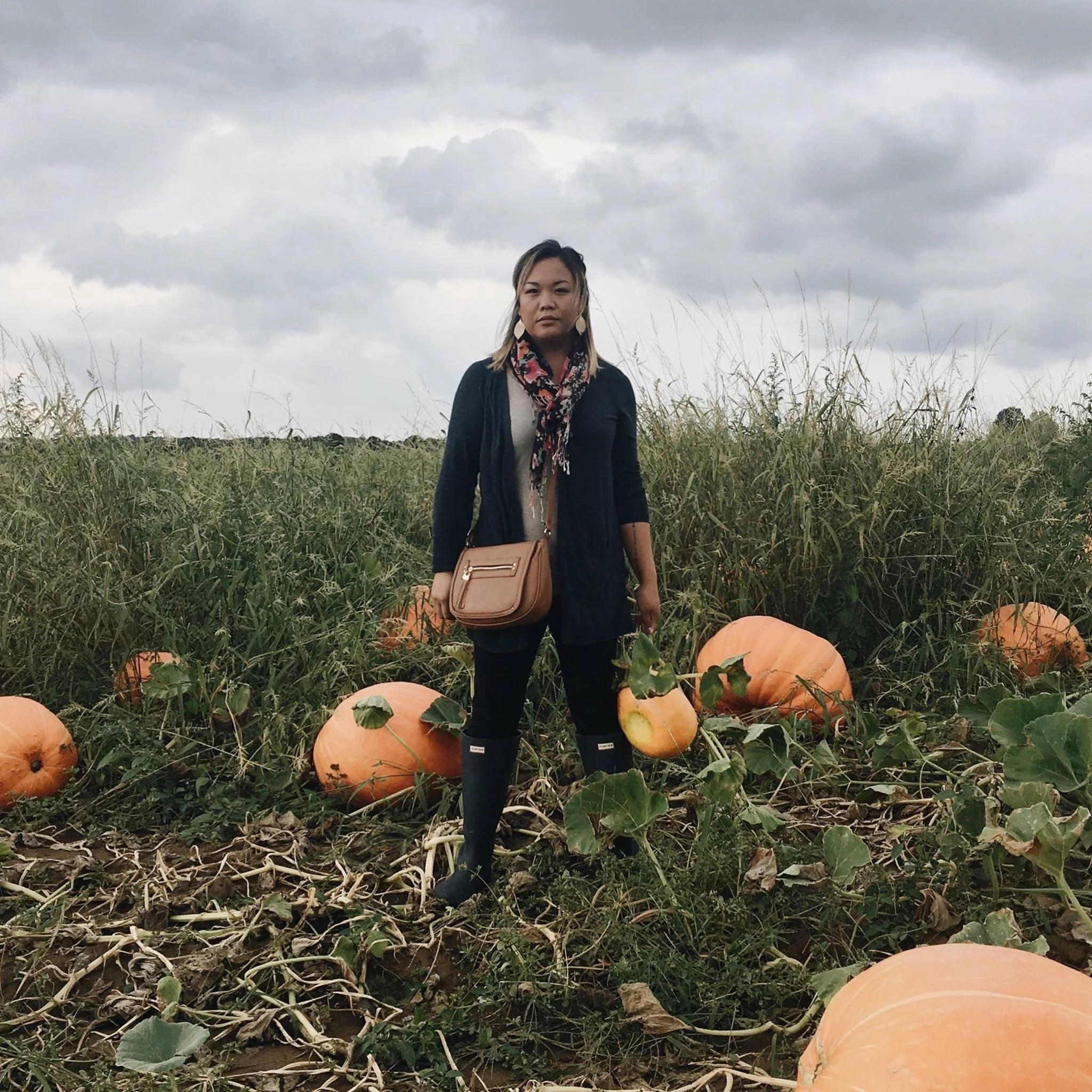 A woman standing in a pumpkin patch with orange pumpkins around her, overcast sky in the background.