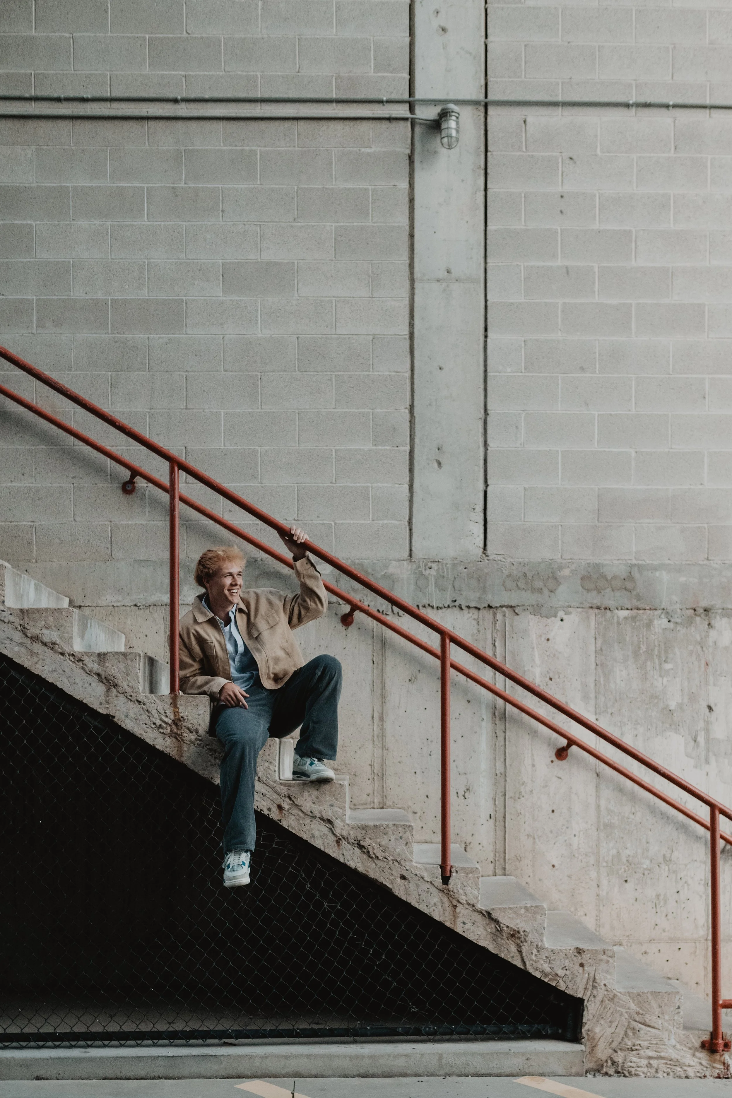 A young man with curly blonde hair sitting on concrete stairs, smiling, wearing a beige jacket, light blue shirt, dark jeans, and sneakers, waving with his right hand. The stairs are beside a concrete wall with a red safety railing.