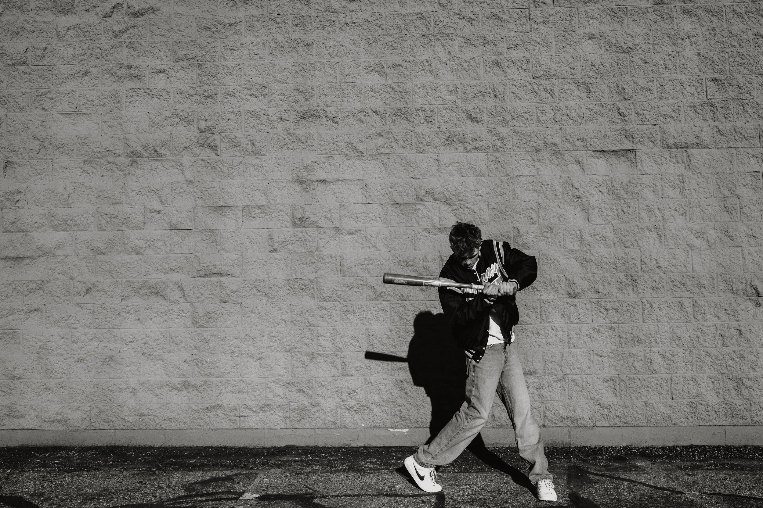 A young man with dark, curly hair wearing a varsity jacket, jeans, and sneakers playing baseball with a bat in front of a large brick wall.